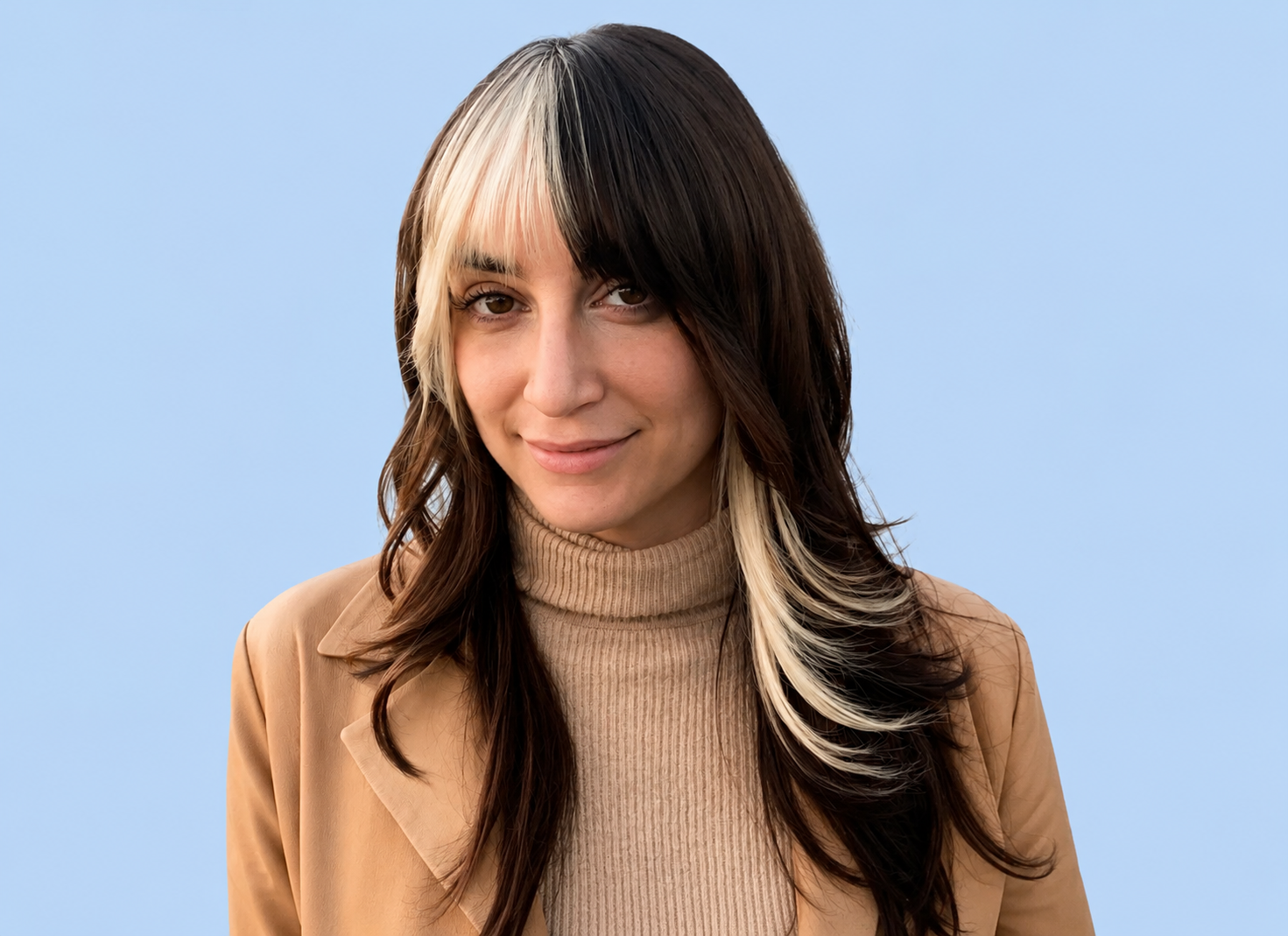 Close-up of a woman with shoulder-length dark hair with blonde highlights, wearing a tan blazer and a beige turtleneck sweater, standing outdoors near a white building with a window, palm trees, and a street visible in the background.