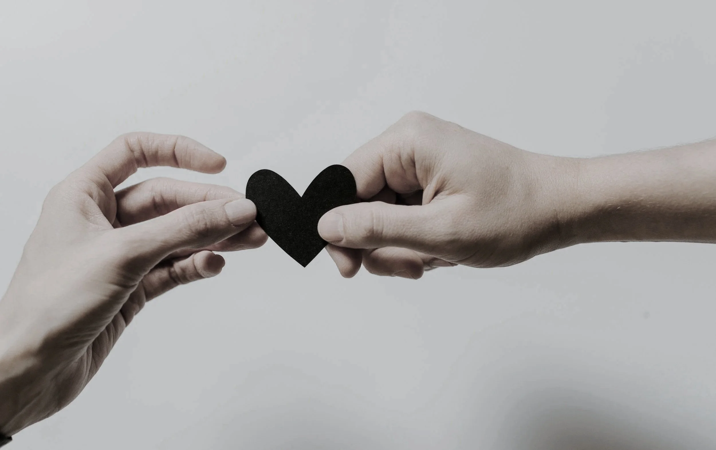 Two hands exchanging a black heart cutout against a plain background.