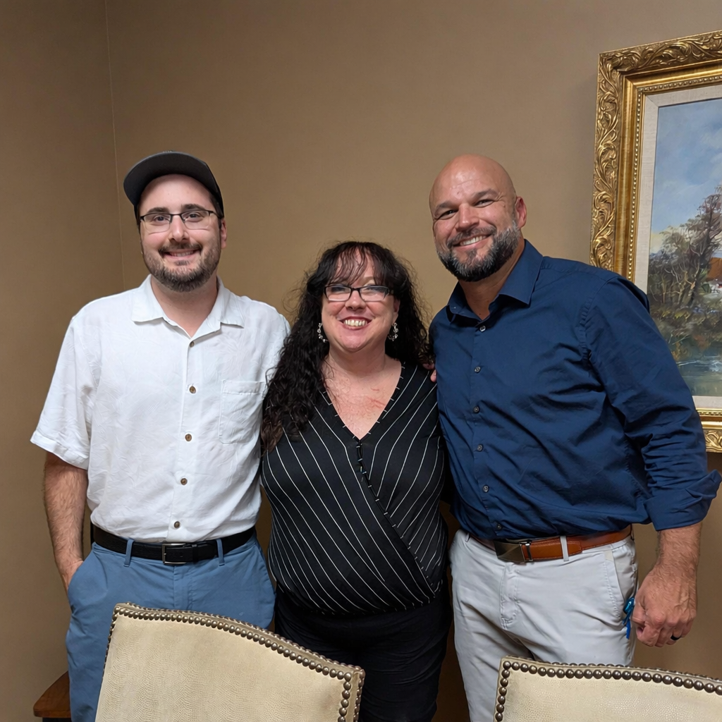 Three people standing together, smiling, in a room with beige walls and a framed landscape painting behind them.