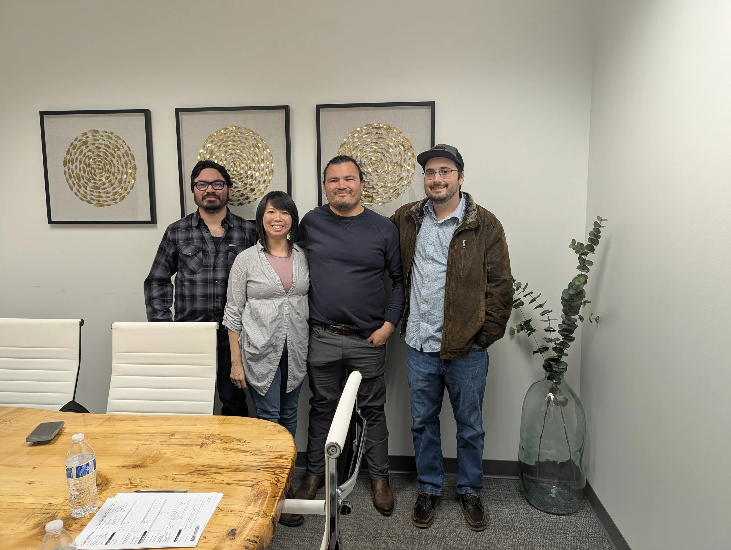 Four people standing in a conference room with white chairs, a wooden table, and abstract gold art on the wall.