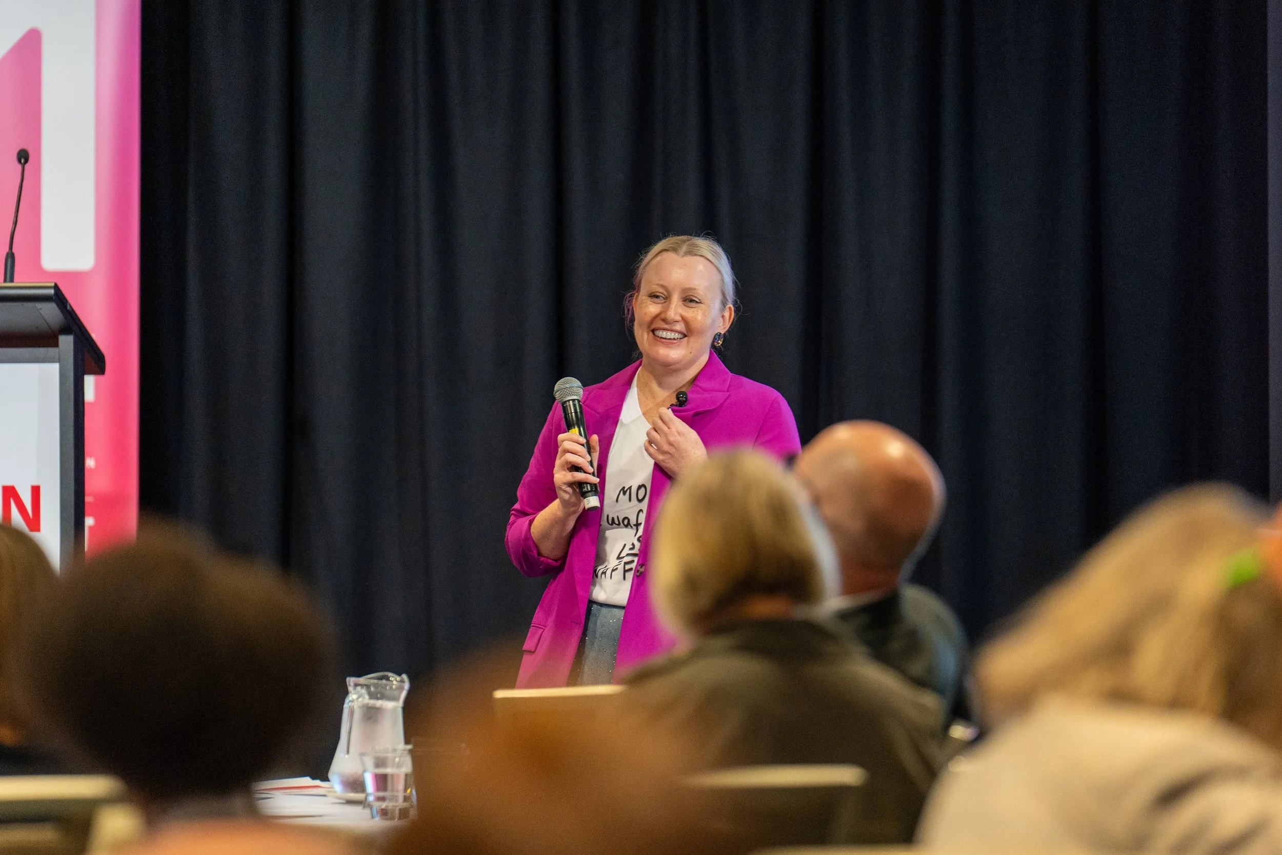 A woman in a pink blazer and white shirt is speaking at an event while holding a microphone, with a dark curtain behind her and an audience in front.