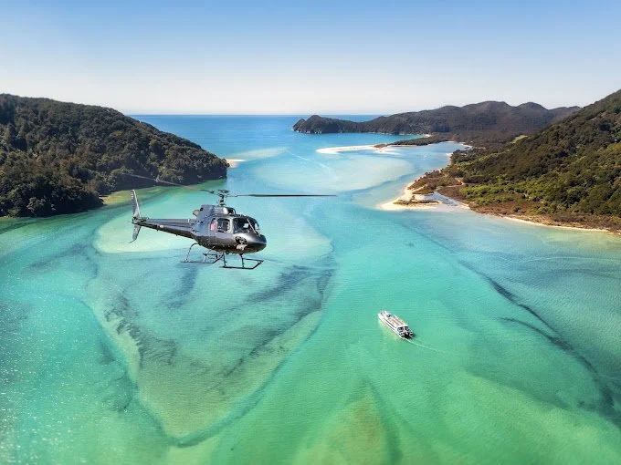 Helicopter flying over turquoise coastal waters and forested hills near Portage Resort, offering scenic flights across Marlborough Sounds, New Zealand.