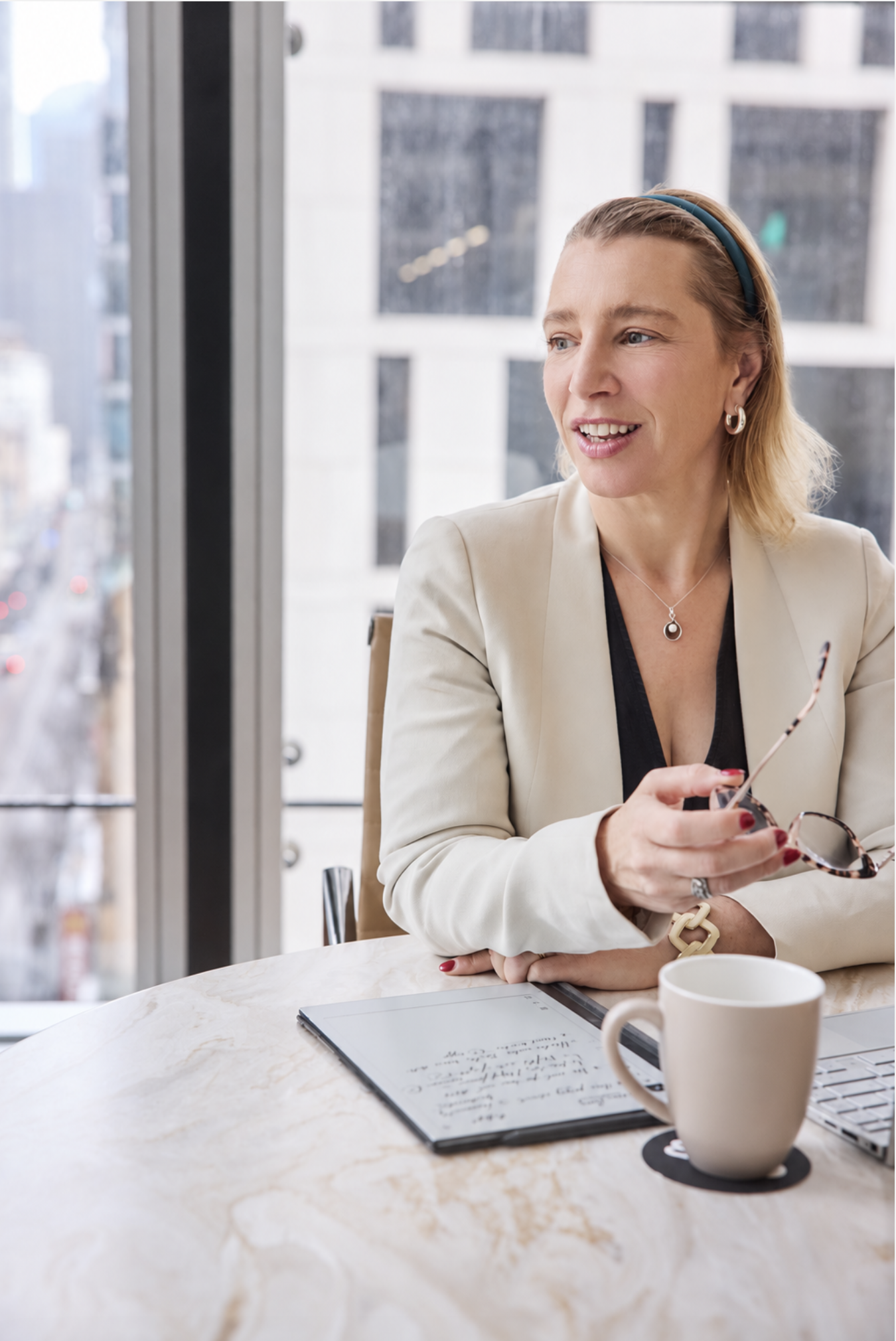 A woman with blonde hair, wearing a cream blazer and silver jewelry, sitting at a marble table with a tablet, a coffee mug, and a notebook. She is holding glasses and looking to her right, with city buildings visible through large windows behind her.
