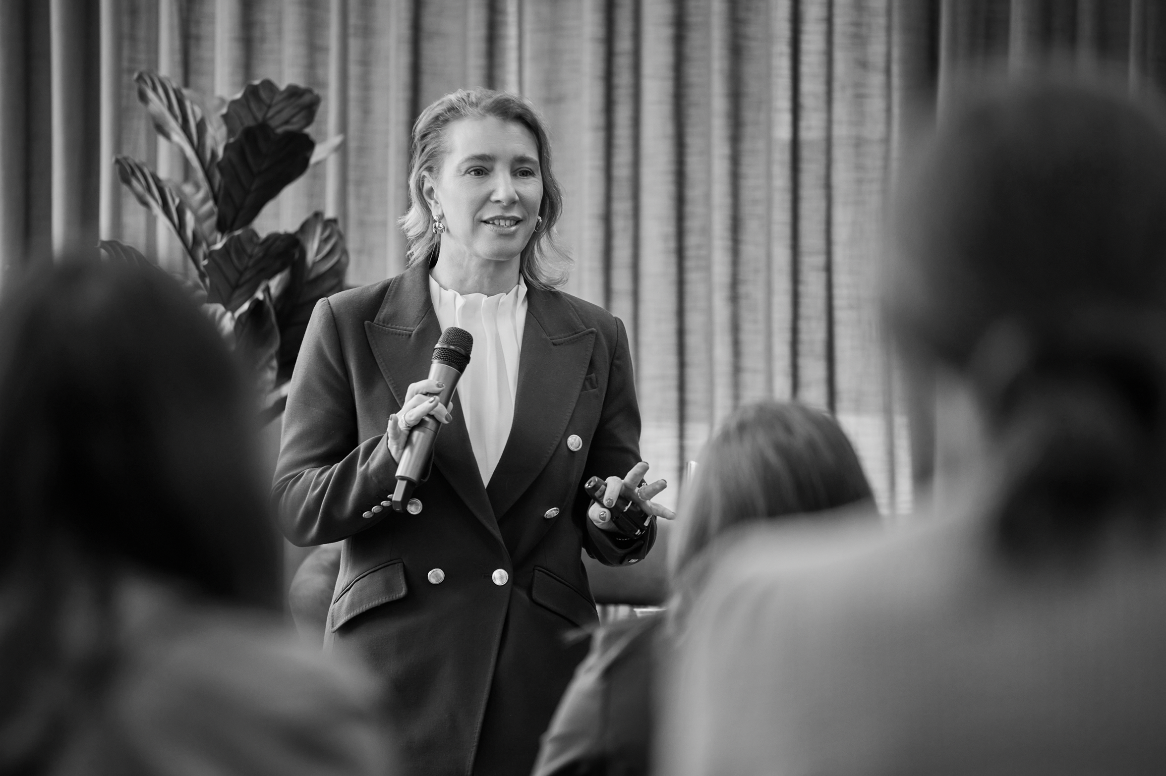 A woman in business attire holding a microphone, speaking to an audience in a professional setting.