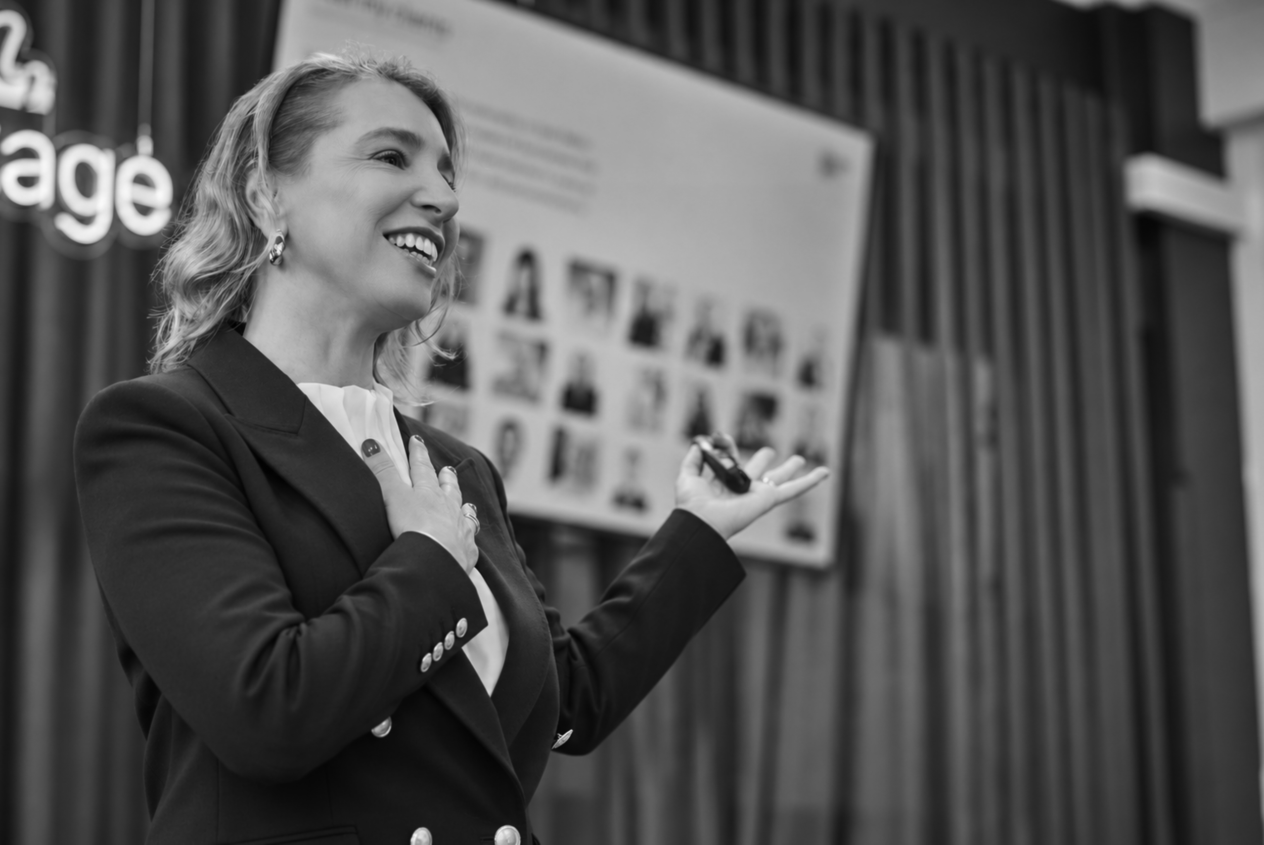 A woman in a suit smiling and speaking, holding a microphone in her right hand, with her left hand over her chest, standing in front of a display board with photos on it.