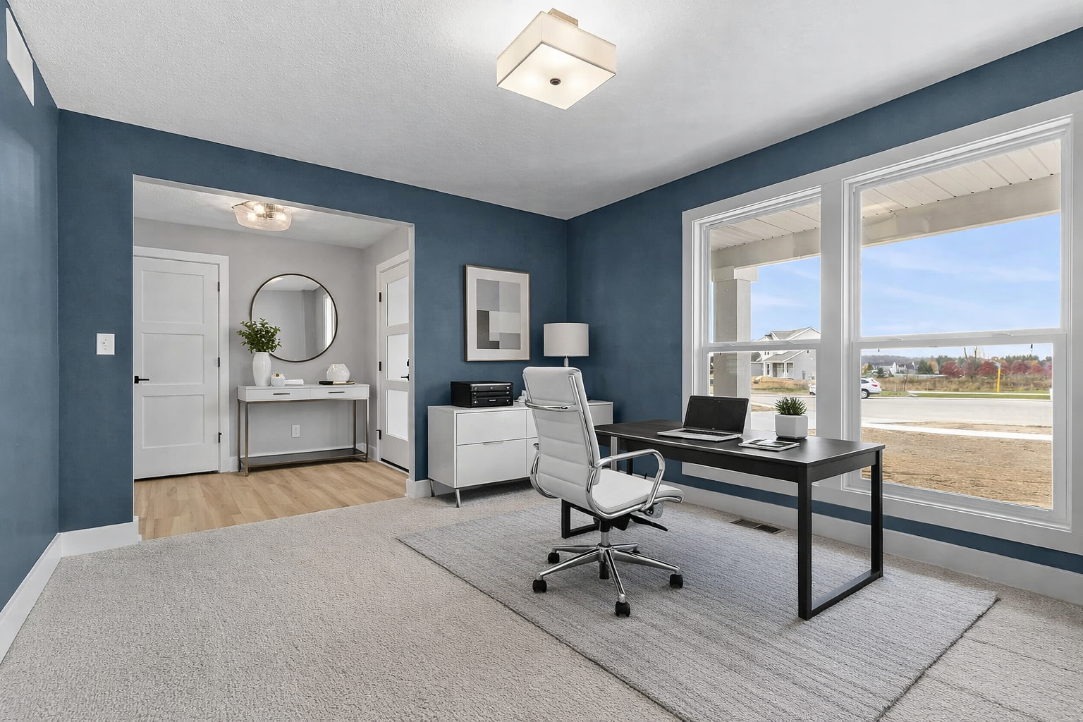 Home office with blue walls, large window showing a street view, black desk with a laptop, white chair, white sideboard, and decor.