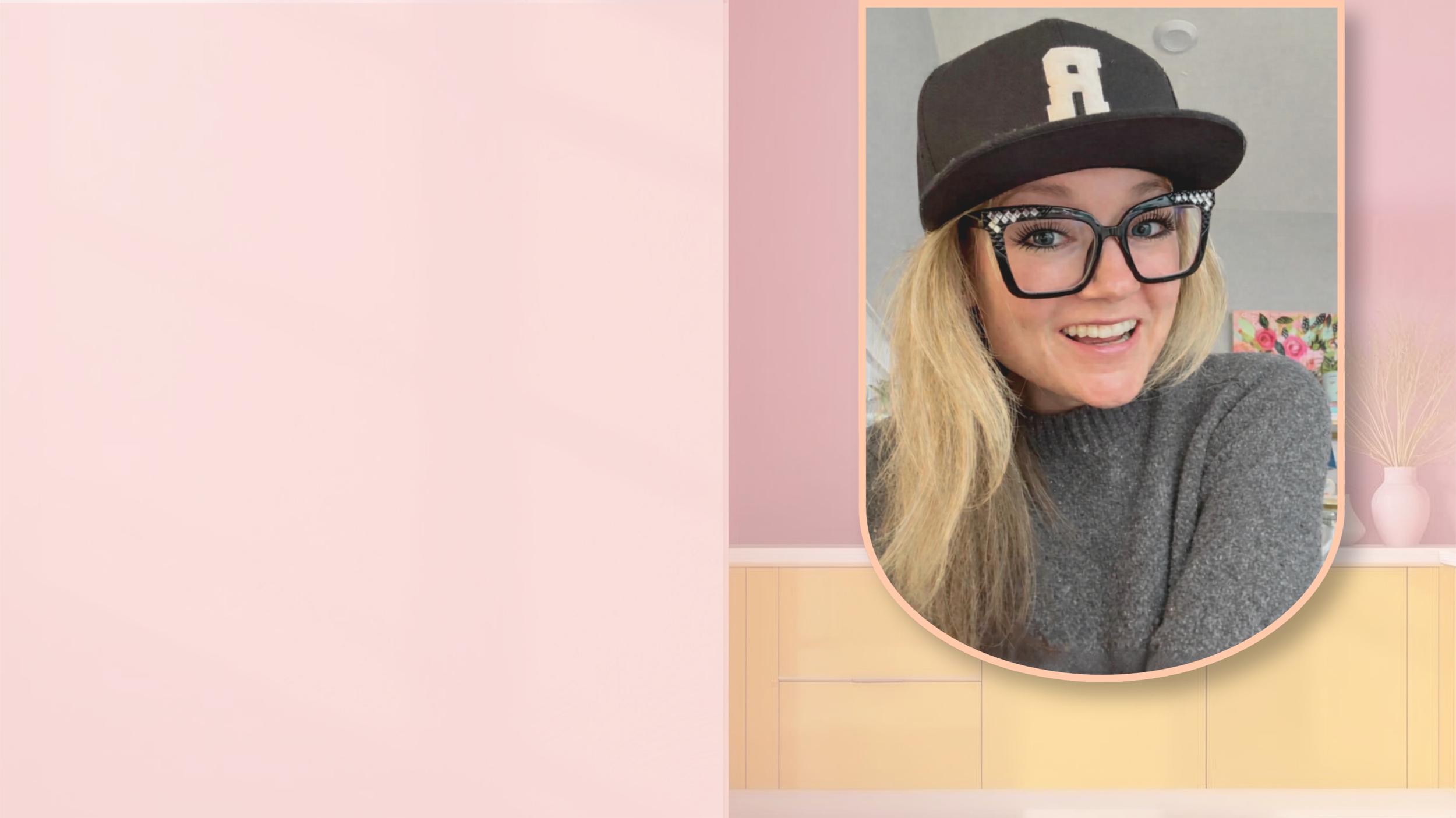 A woman with blonde hair wearing large glasses, a black baseball cap, and a gray sweater, smiling in front of a pink and yellow background with a vase of pampas grass.