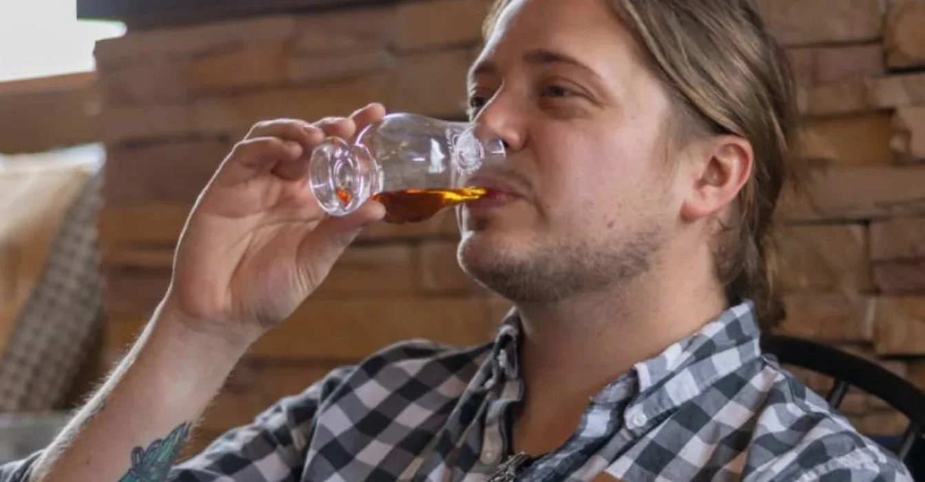 White man in a blue and white checked shirt drinking whiskey from a glencairn glass