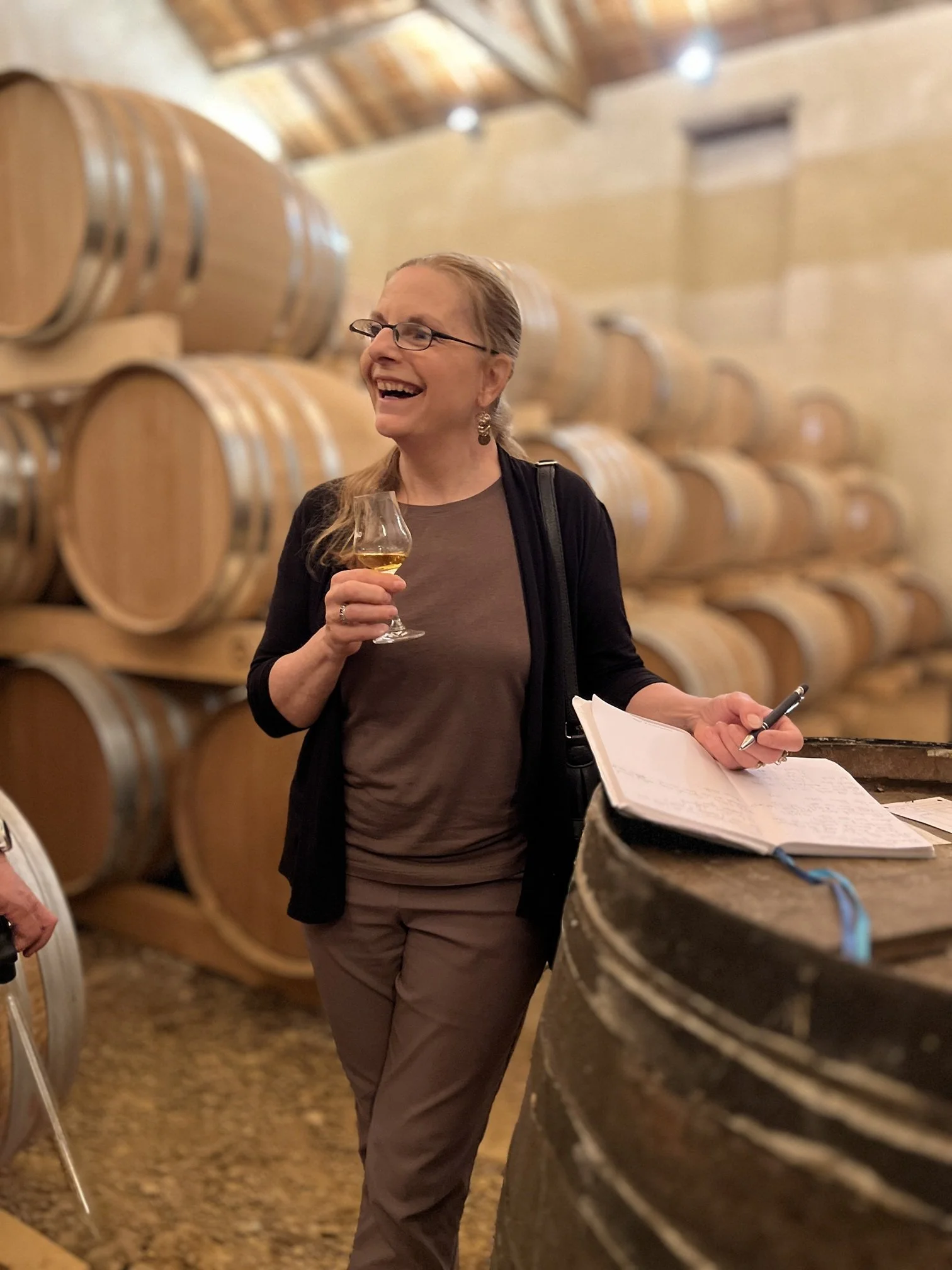 A smiling woman wearing glasses stands in a Cognac or Armagnac aging cellar filled with stacked oak barrels, holding a glass of spirit and a notebook with a pen.