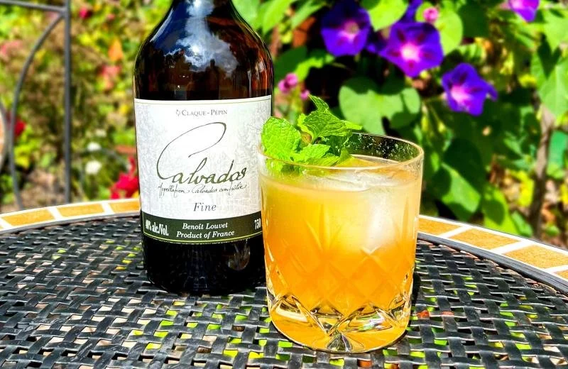 A rocks glass with a yellow-orange cocktail garnished with mint leaves, placed next to a bottle of Claque-Pépin Calvados Fine on a black metal outdoor table, with purple and pink flowering plants in the background.