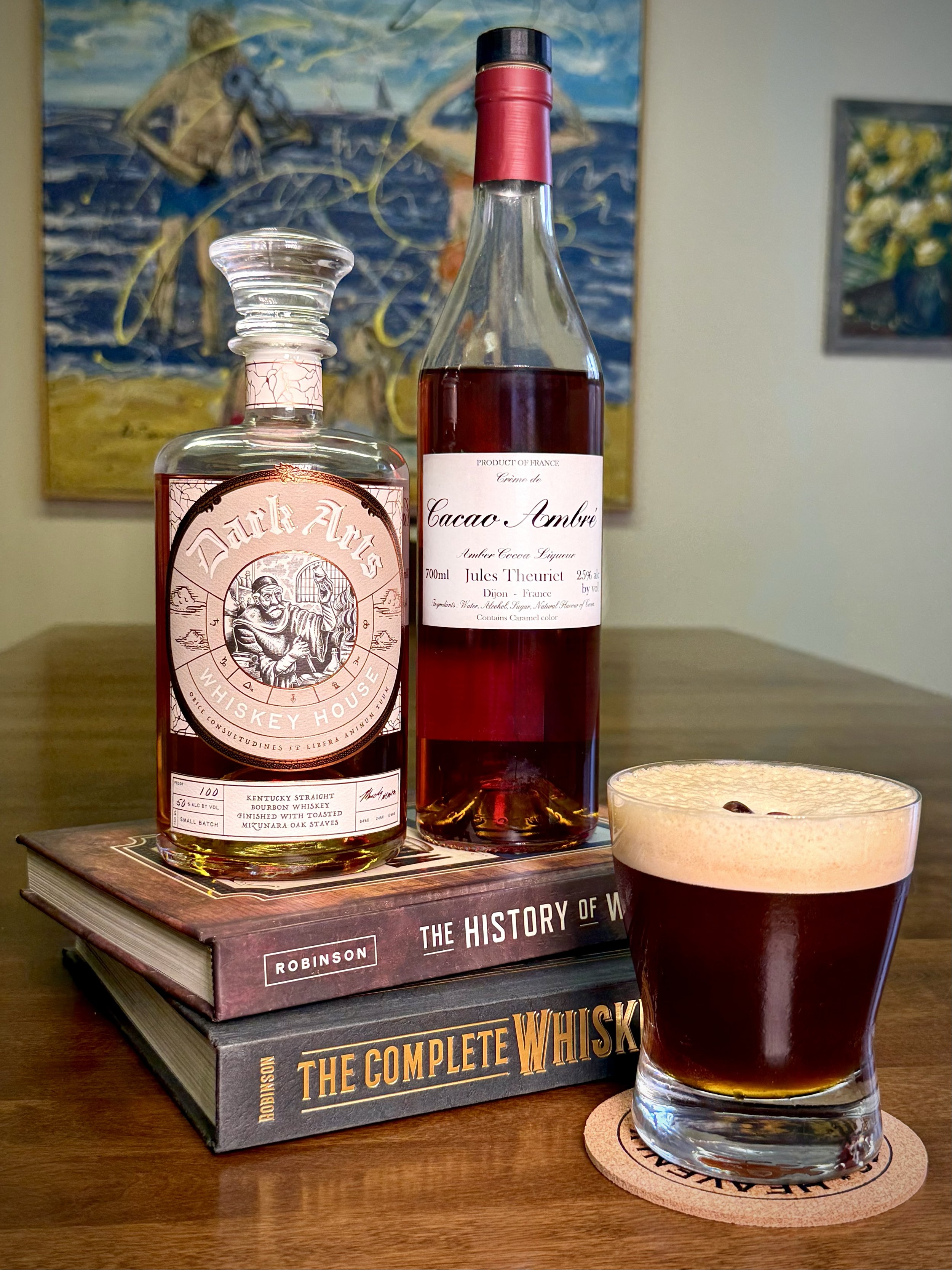 Bottles of Dark Arts Whiskey and Jules Theuriet Cacao Ambré rest on whiskey books next to a dark, frothy cocktail topped with coffee beans. A landscape painting hangs in the blurred background.