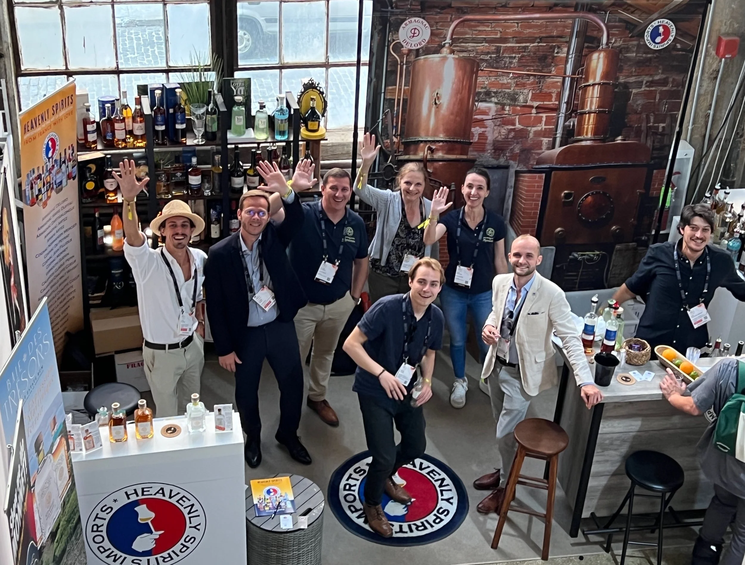 Group of eight people looking upwards, smiling and waving at an exhibition booth for Heavenly Spirits, with a bar setup and bottles of alcohol displayed behind them.