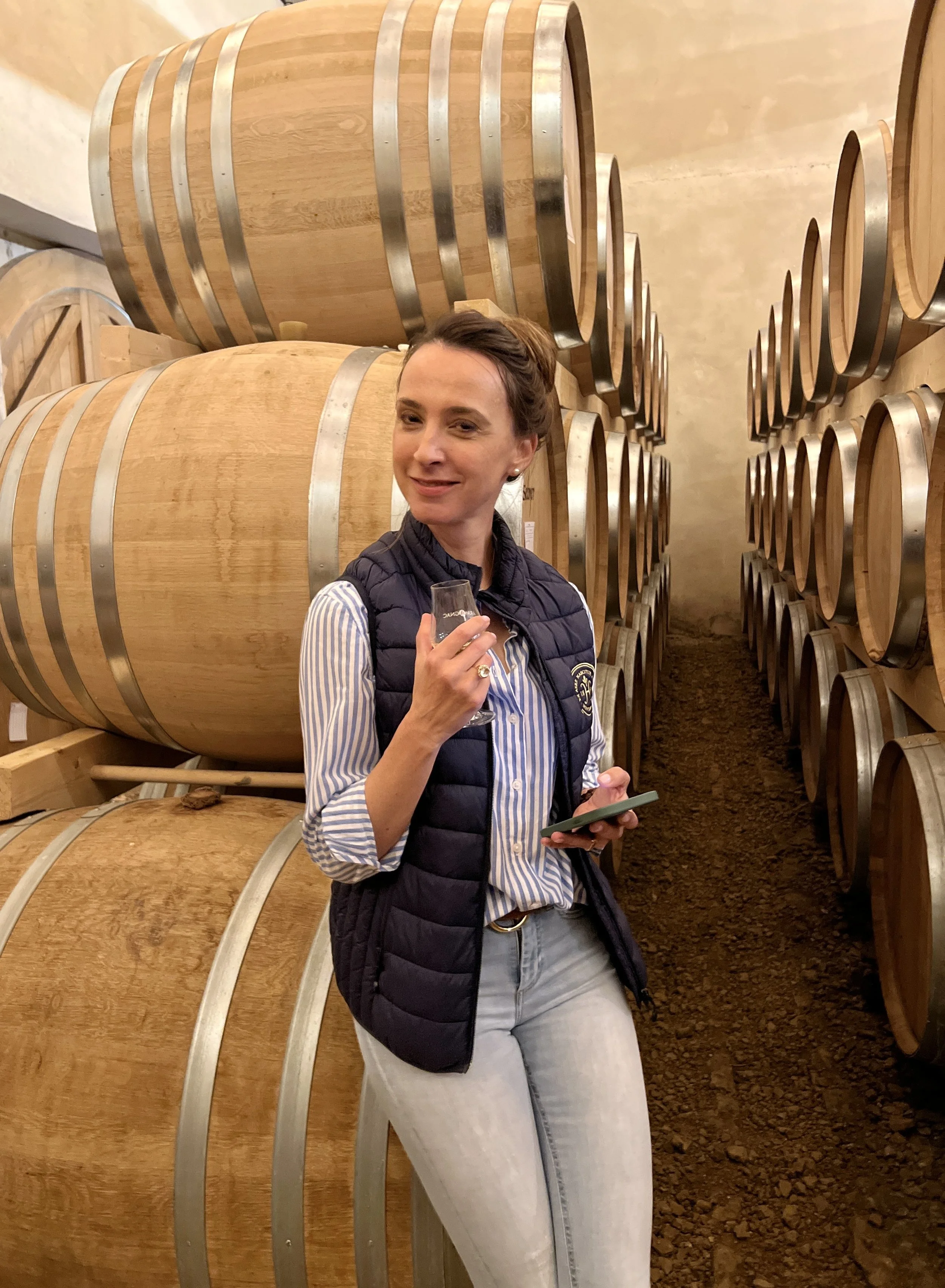 Cindy Crighton leans against a stack of Armagnac barrels in the cellar of Château de Hontambère, holding a tasting glass and smiling at the viewer.