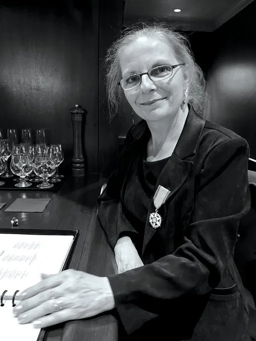 Black and white photo of a woman in glasses sitting at a bar wearing a medal attached to her blazer