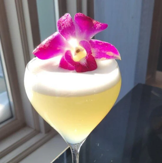 Pale yellow cockail in long stemmed glass with white froth garnished with a fuchsia orchid