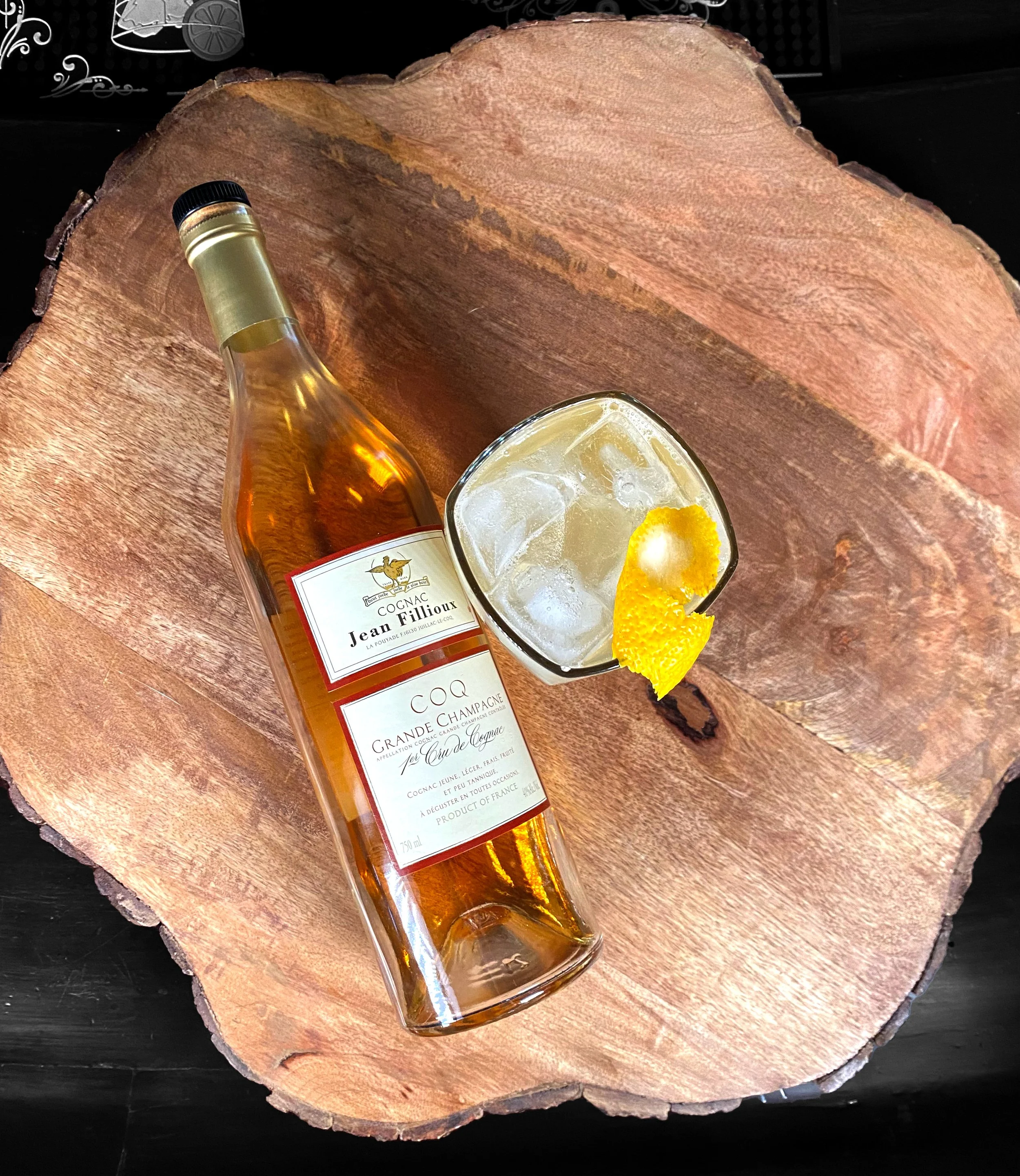 Bottle of Jean Fillioux Cognac Coq lying down next to a top-down view of a cocktail with ice and lemon peel garnish