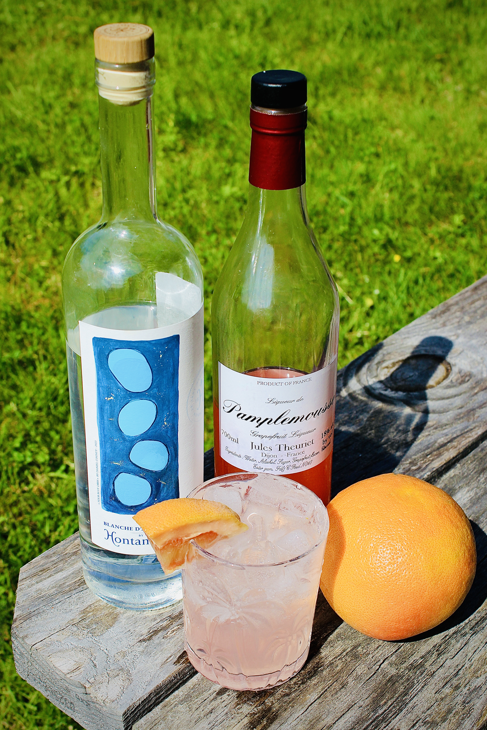A pale pink French Paloma cocktail in a cut glass rocks glass garnished with a grapefruit wedge with bottles of Hontambère Blanche Armagnac and Jules Theuriet Pamplemousse liqueur behind it next to a whole grapefruit on wooden picnic table outside.