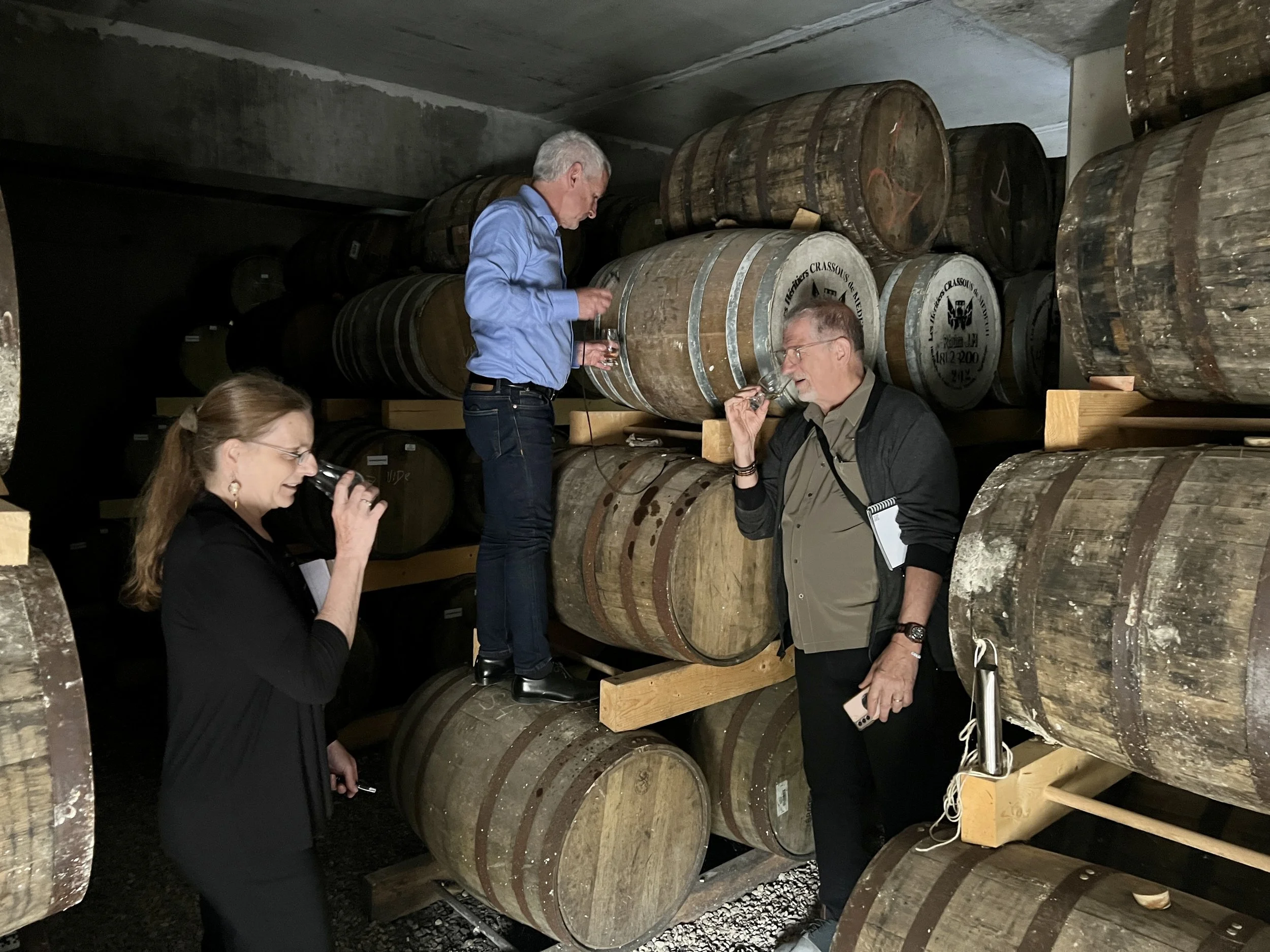Three people tasting whiskey in a cellar with stacked barrels.