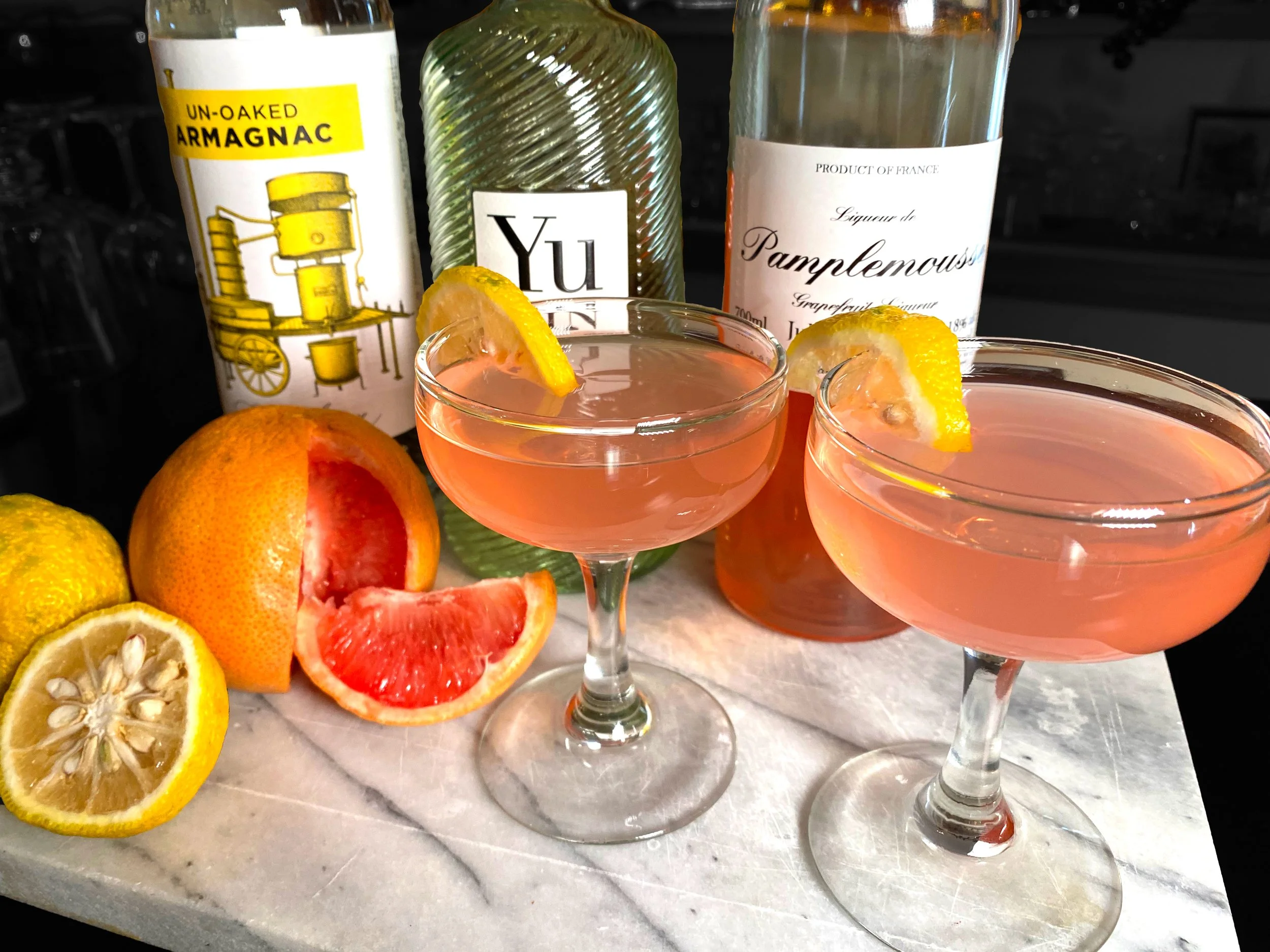 Two pink cocktails in coupe glasses with lemons, grapefruit, and bottles in the background