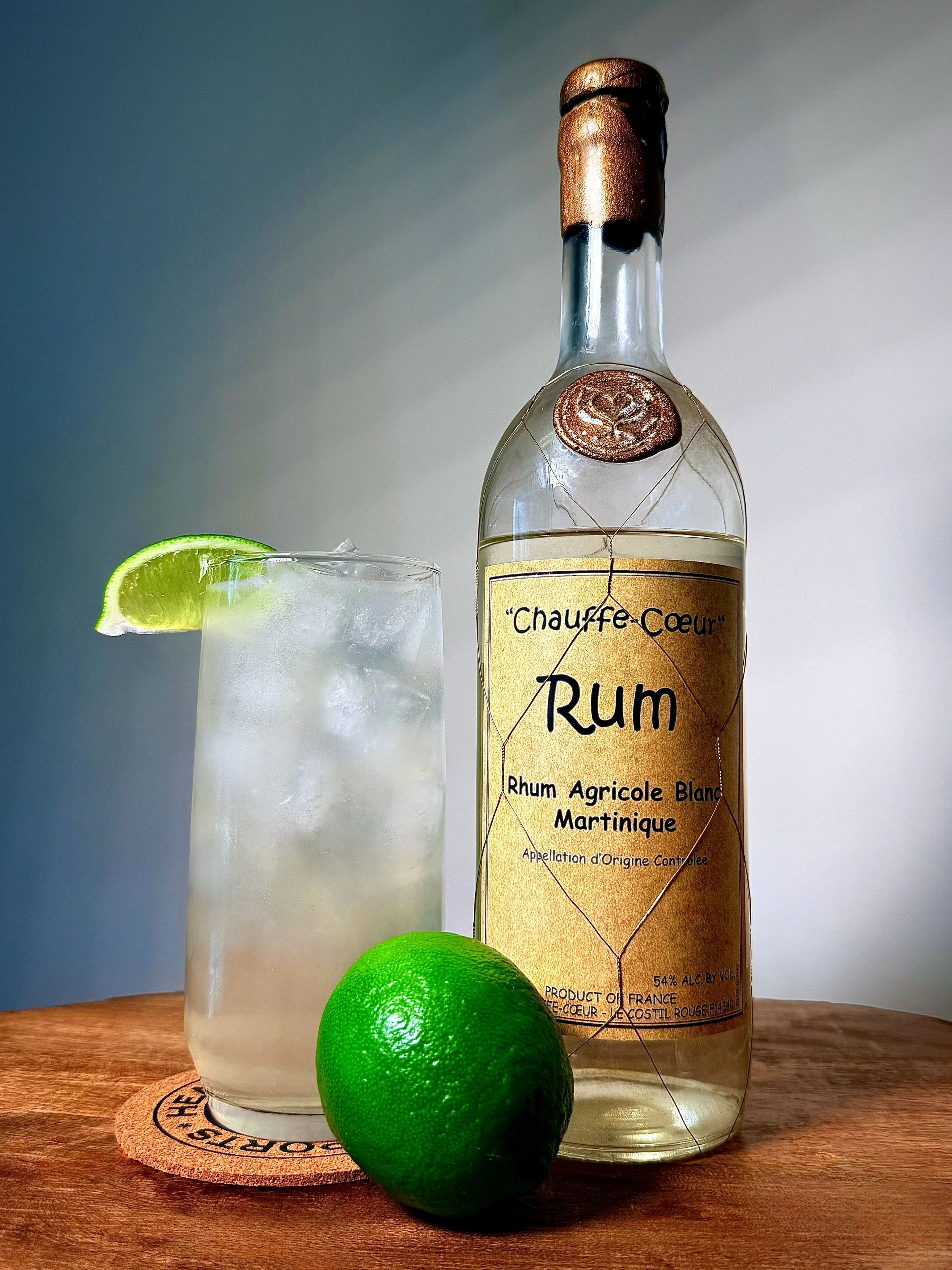 A vibrant Bright & Stormy CC cocktail in a tall glass, garnished with a lime wedge and mint, next to a bottle of Chauffe-Cœur Rum Agricole Blanc from Martinique with a copper wax seal.