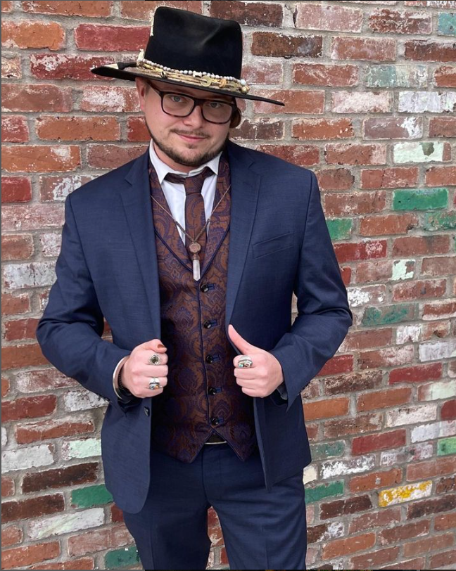 White man in decorated fedora, blue suit and brown vest poses in front of a brick wall