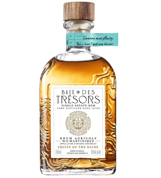 A  square-based bottle of Baie des Trésors single estate Rhum Agricole Martinique Fruits of the Rains with a wooden cap and a white label featuring black text and gold emblem, filled with amber-colored rhum, aged in small French oak barrels.