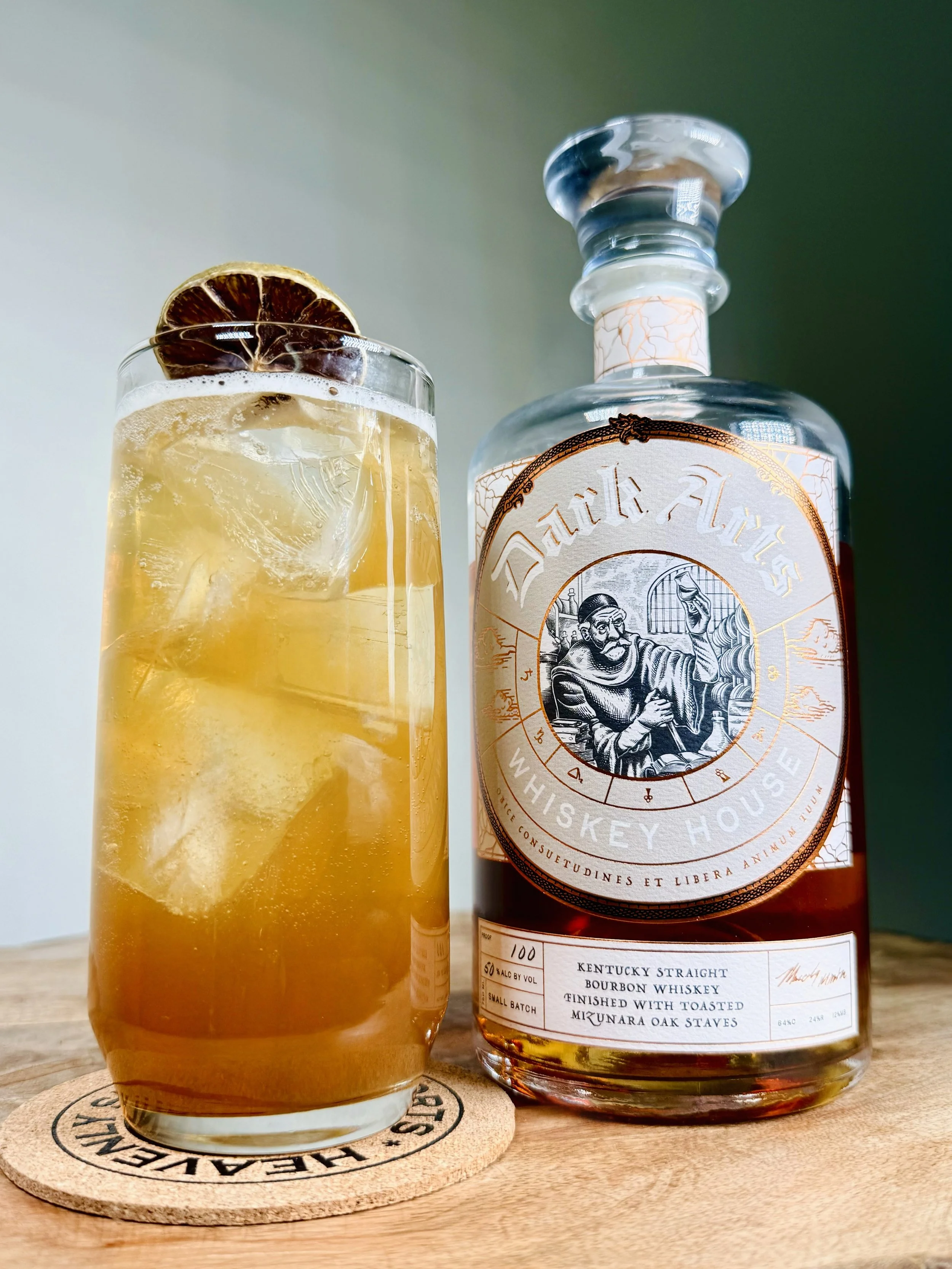 Maple Bourbon Rickey cocktail with a dried lime garnish, featuring a bottle of Dark Arts Whiskey. Refreshing tall bourbon cocktail  for craft whiskey enthusiasts.