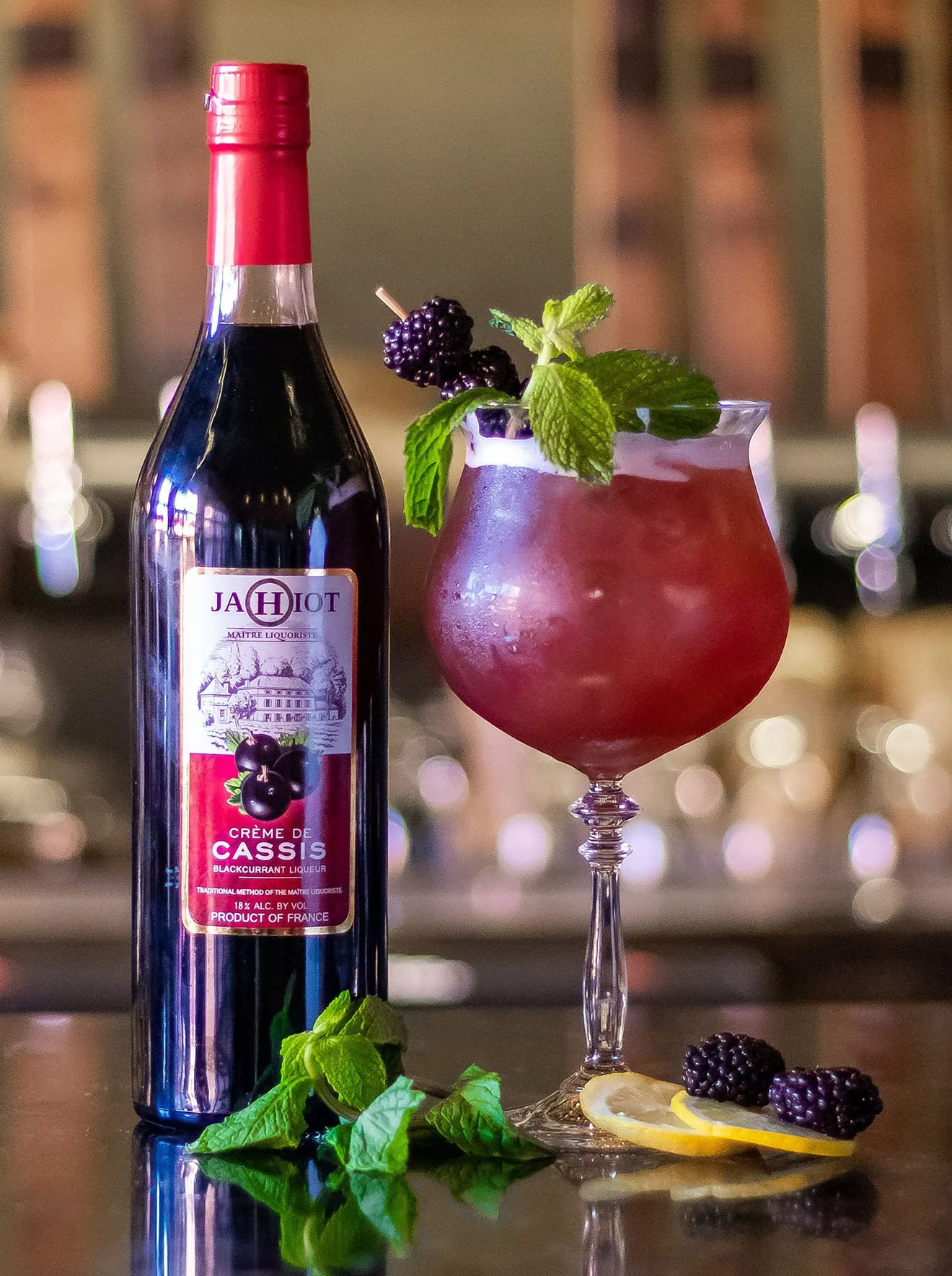Bottle of Jahiot Crème de Cassi next to a cranberry colored cocktail garnished with mint and blackberries