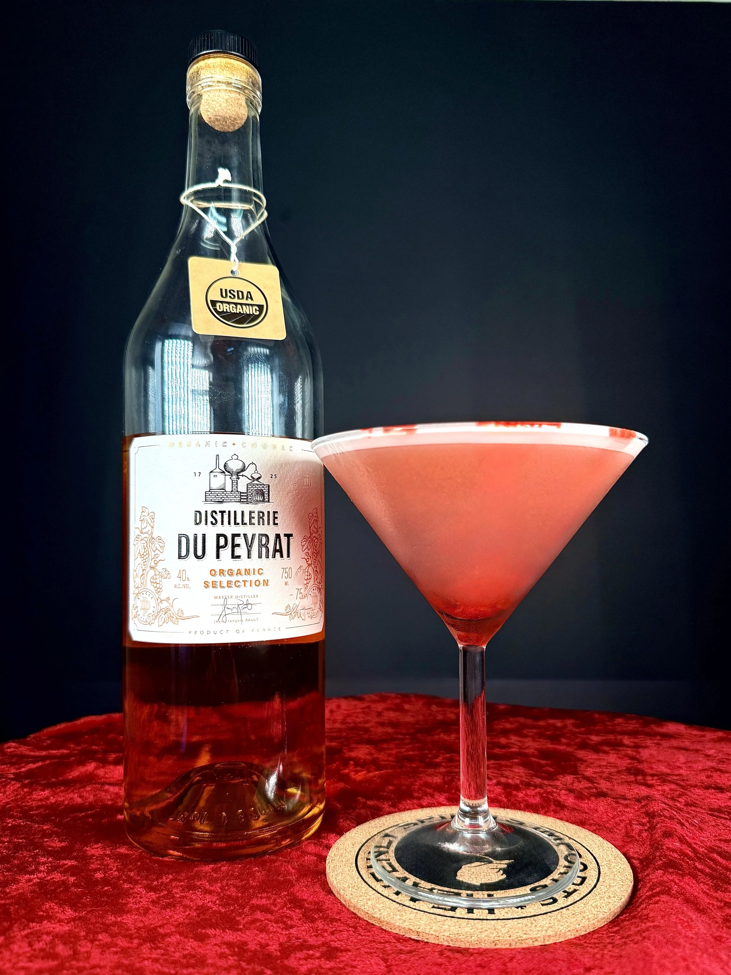 A red layered cocktail in a martini glass is paired with a bottle of Distillerie Du Peyrat Organic Cognac. The set is presented on a red velvet surface against a stark black background