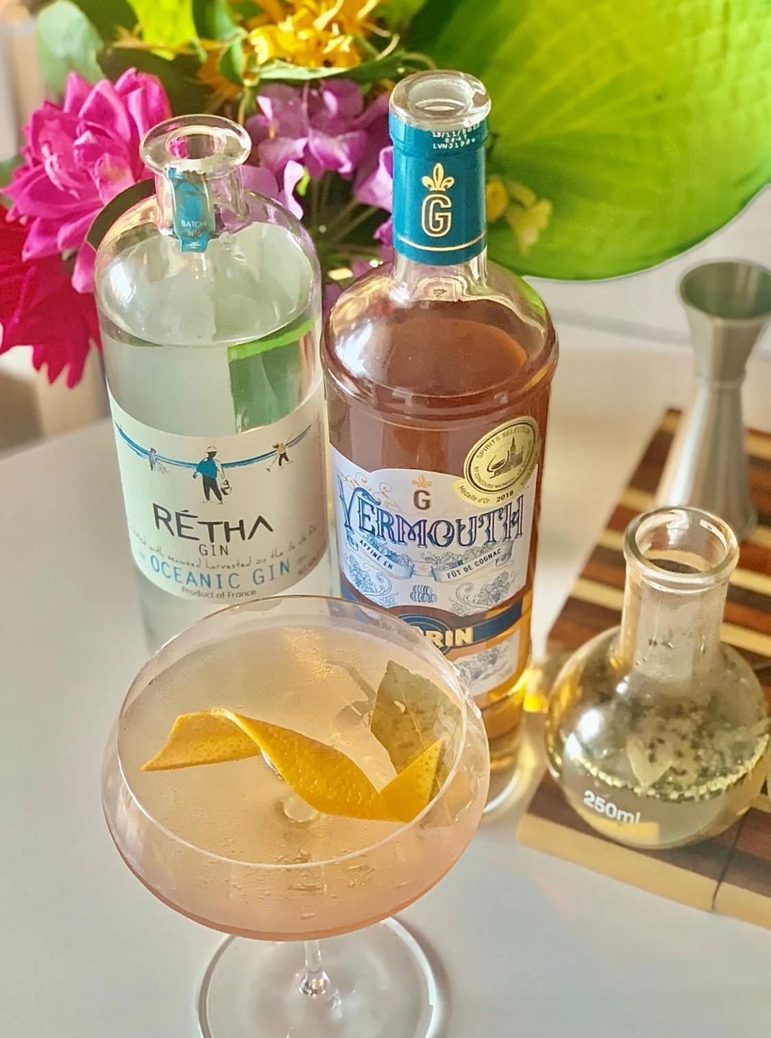 Close up of pale yellow cocktail in a coupe with lemon twist and bay leaf with two bottles and bar tools in the background