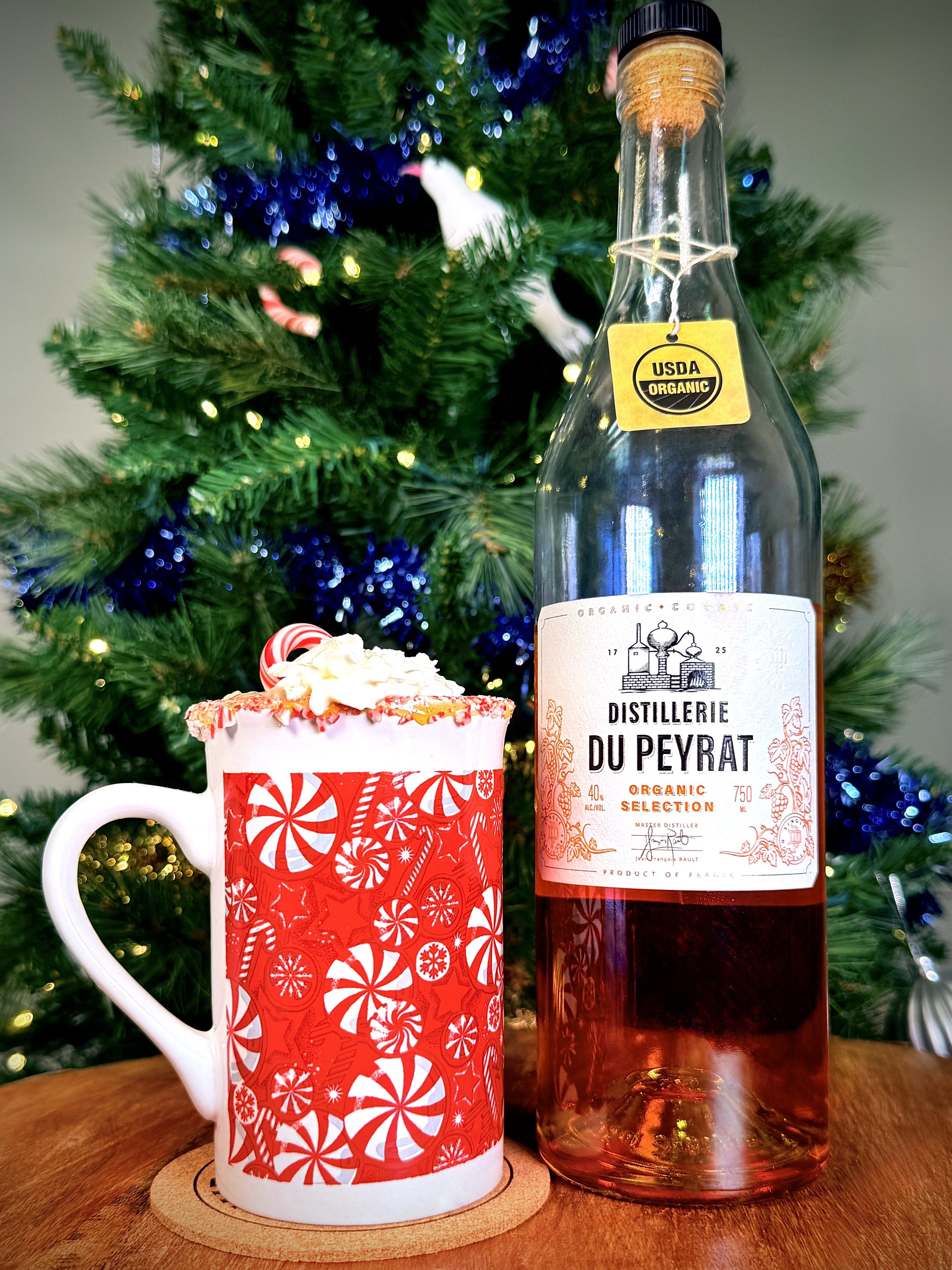 Festive holiday spiked hot cocoa featuring Distillerie Du Peyrat Organic Cognac. Served in a peppermint-themed mug with whipped cream and a candy cane in front of a Christmas tree.