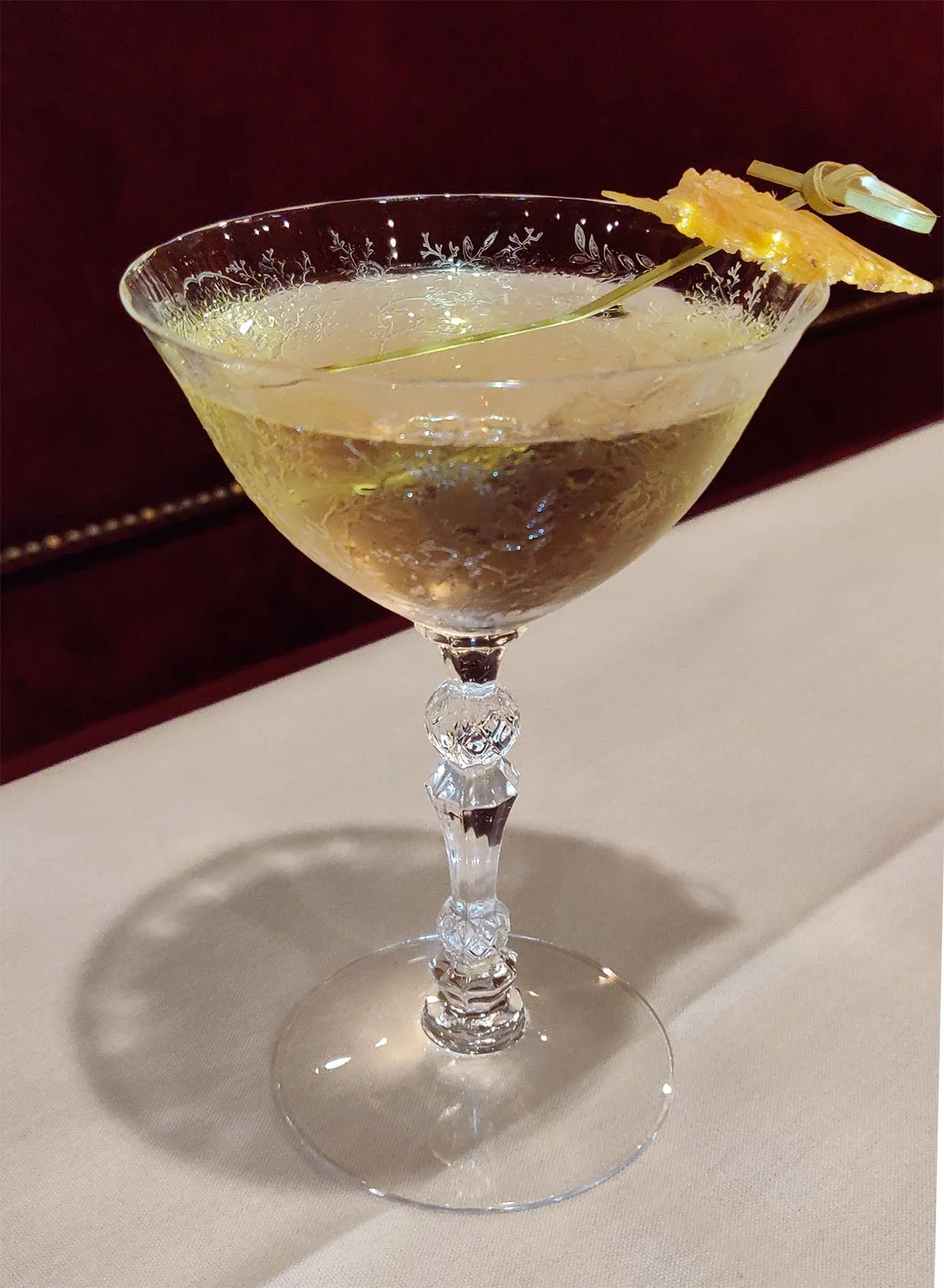 Pale yellow cocktail in a fancy crystal glass with orange peel garnish on a bamboo cocktail pick