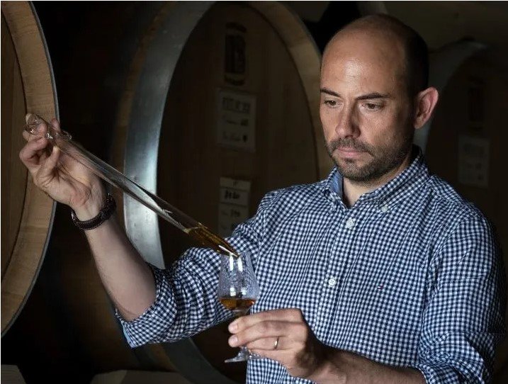 Man in a blue checked shirt measures Armagnac into a glass in front of a barrel