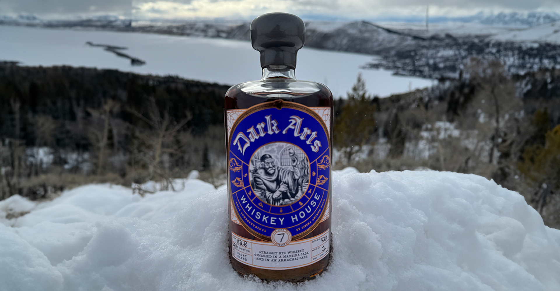 Blue labeled bottle of whisky sitting in the snow overlooking a landscape of a lake and mountains