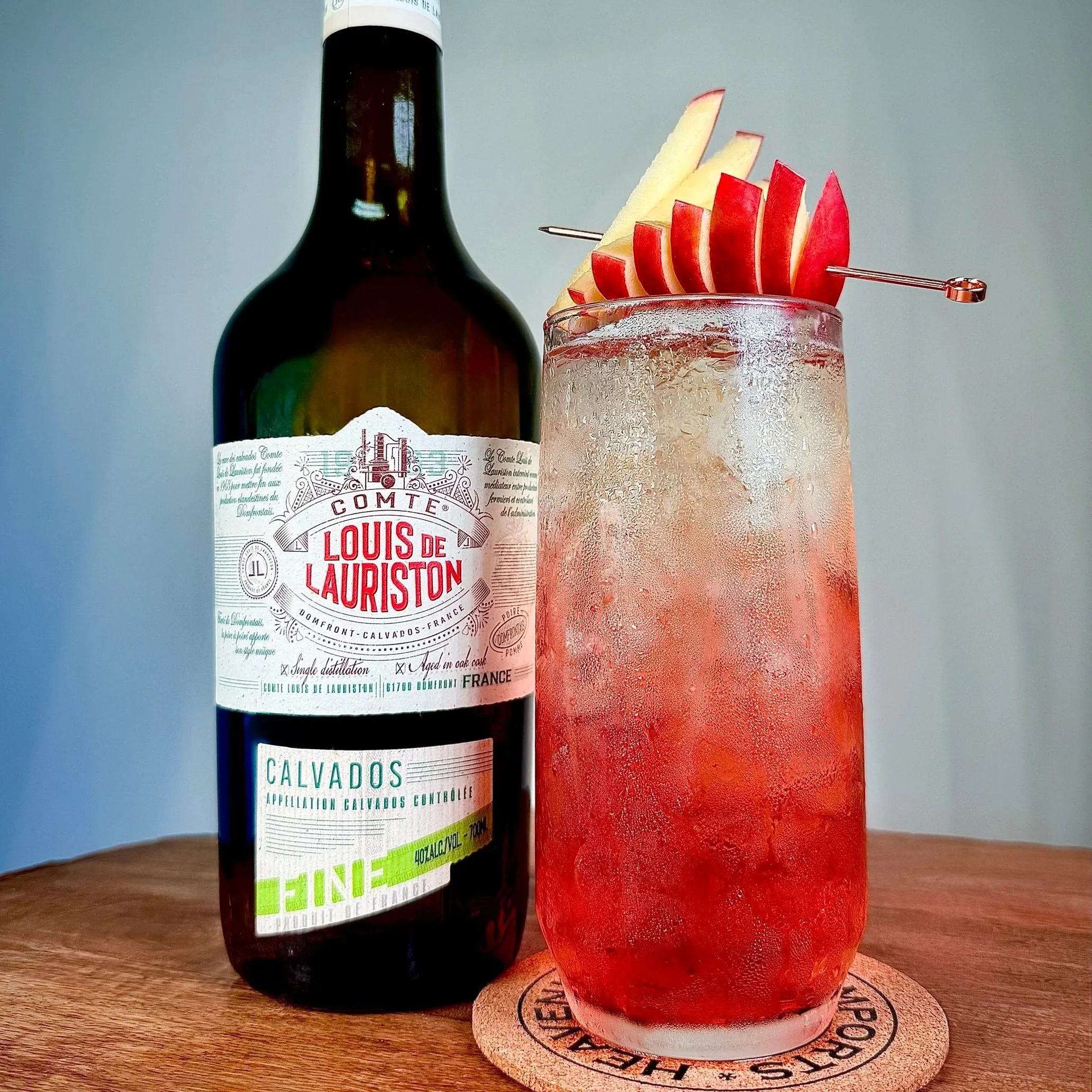 Comte Louis de Lauriston Calvados bottle next to a vibrant Calva Cranberry cocktail in a highball glass, garnished with sliced apple, on a wood surface and cork coaster.