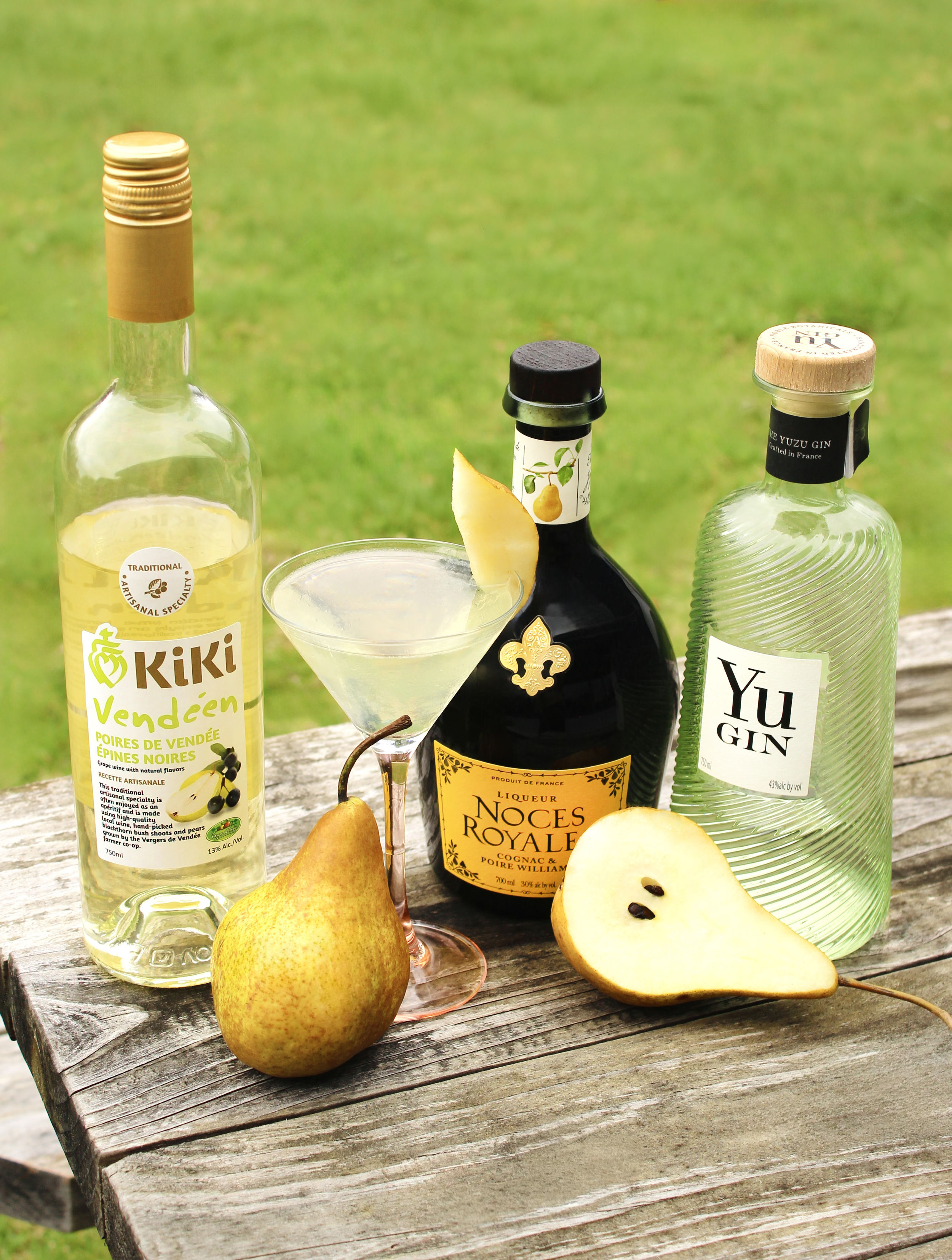 A martini glass filled with a clear cocktail and a pear slice sits on a rustic wood table. Behind it are bottles of KiKi Pear Vendéen Apéritif, Yu Gin, and Noces Royales, accompanied by a whole and a halved pear.