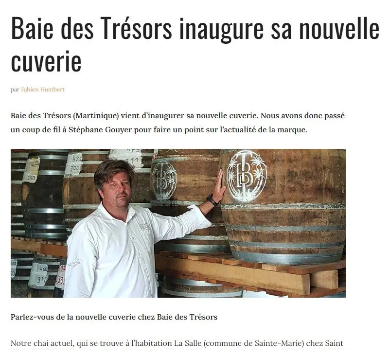 A man in a white button down stands next to large wooden rum barrels inside an aging cellar, touching one of the barrels. The barrels feature a large white logo painted on them featuring a T growing through a B with palm trees on either side