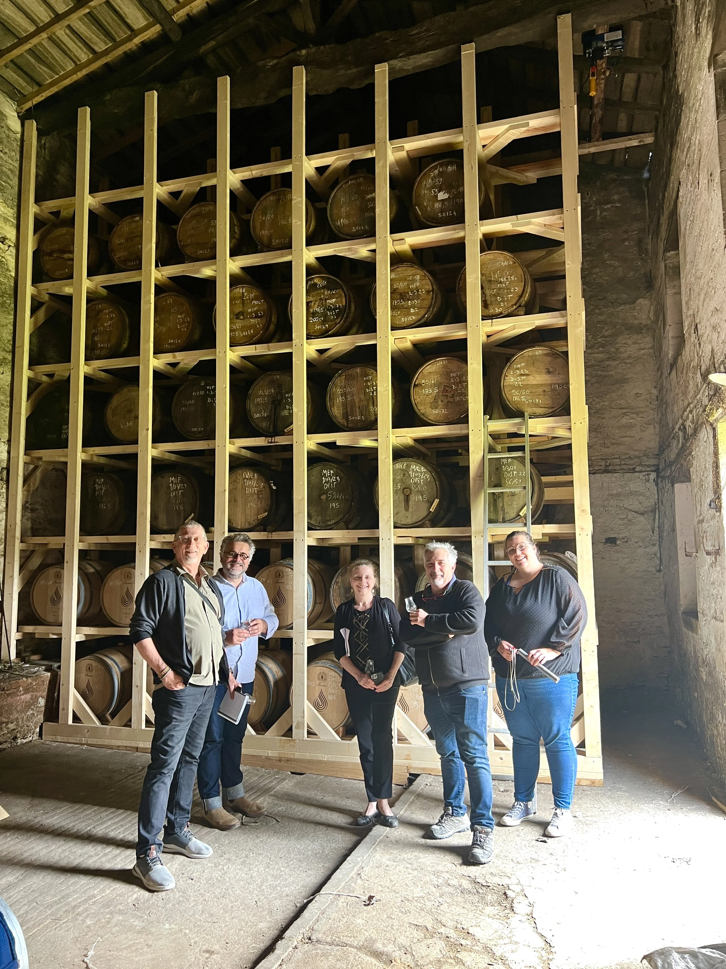 Group of five people standing in front of a large wooden whisky barrel storage rack stacked six layers high with an empty seventh layer inside a rustic cellar.