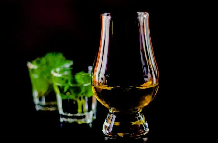 Glencairn glass with whisky with two shot glasses of white spirits and mint out-of-focus behind on black background