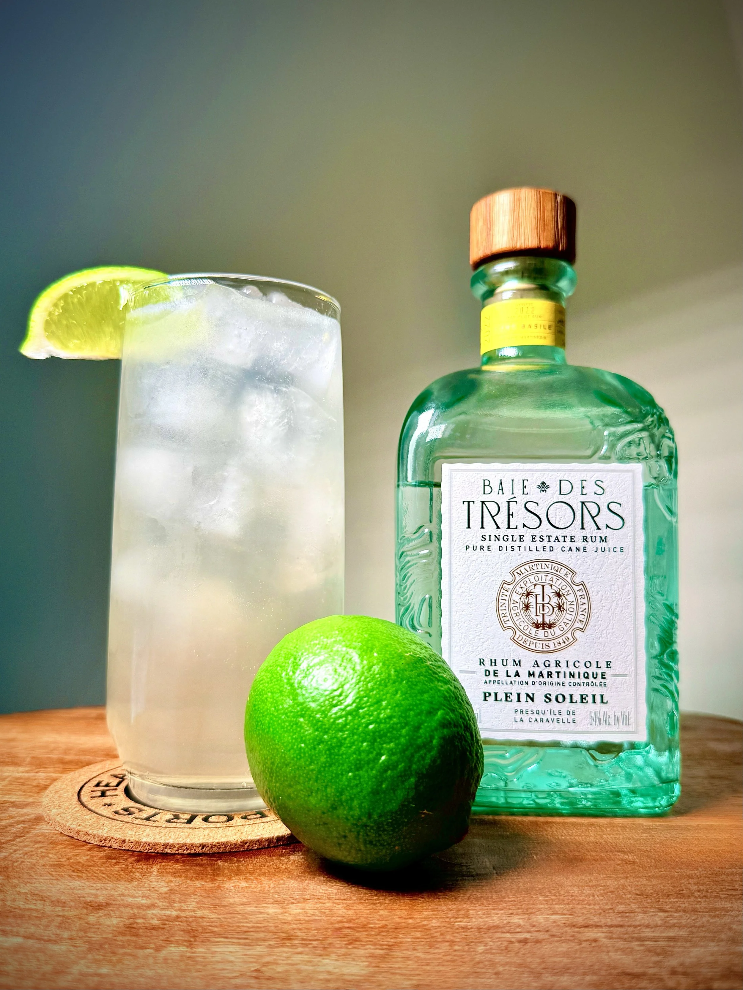 A clear, effervescent Bright & Stormy BDT cocktail in a highball glass, garnished with a lime wedge next to a bottle of Baie des Trésors Rhum Agricole Martinique Blanc "Plein Soleil" in a blue square-bottomed bottle with raised topographical lines.