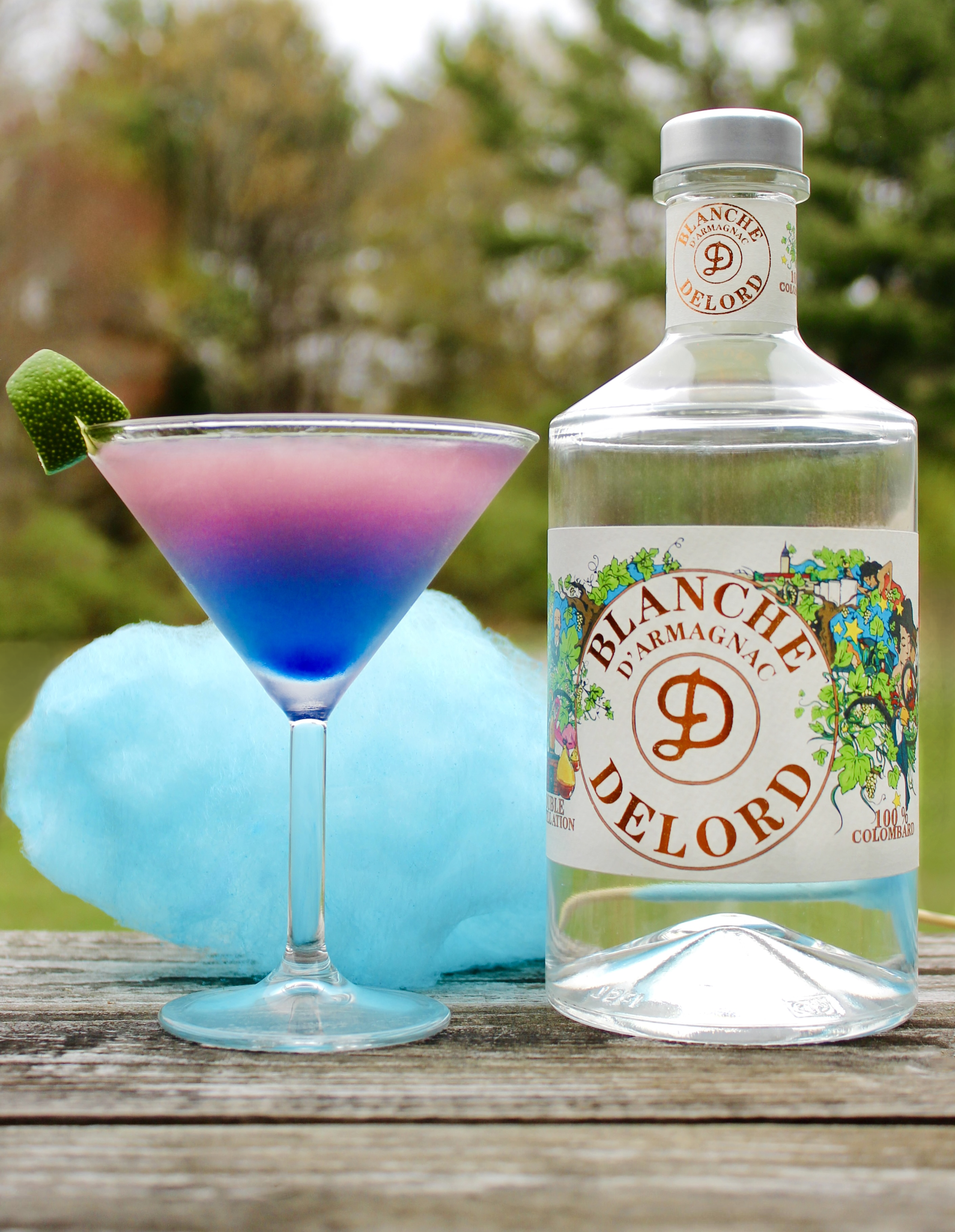 A martini glass displays a vibrant two-toned pink and blue cocktail garnished with lime. A massive tuft of light blue cotton candy rests behind the glass next to a bottle of Delord Blanche Armagnac.