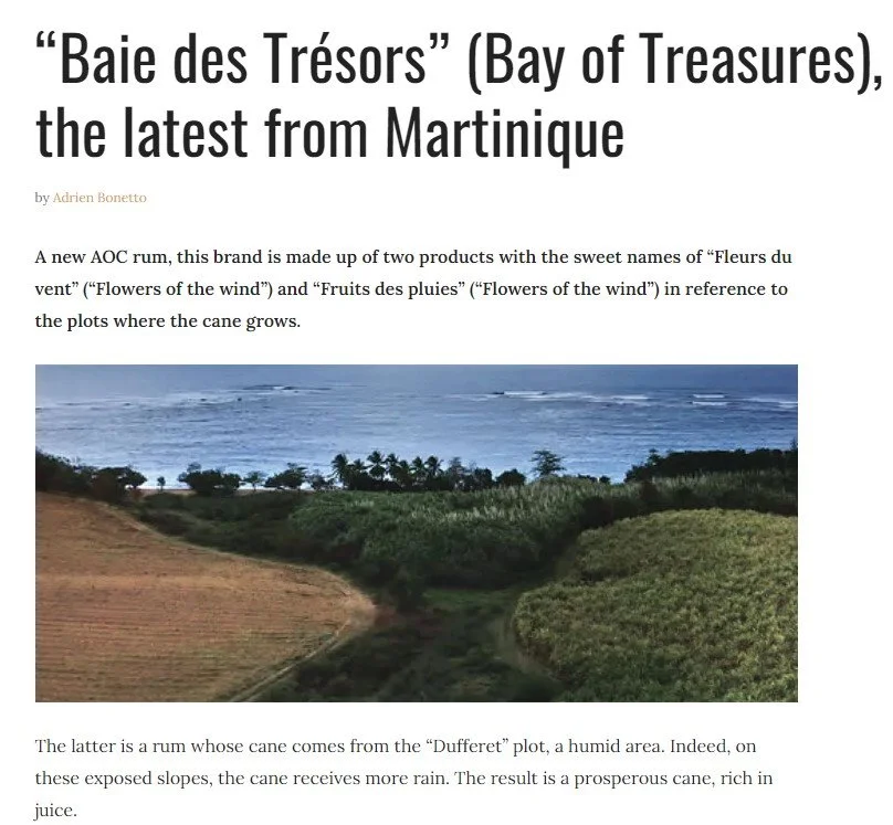 A magazine article featuring a landscape photo of the Bay of Treasures in Martinique, with lush green fields, trees, and the ocean in the background and the headline "'Baie des Trésors' (Bay of Treasures), the latest from Martinique."