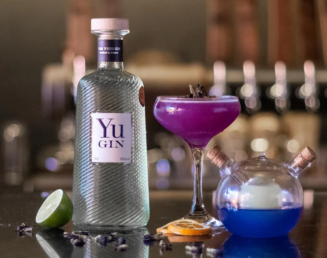 Bottle of Yu Gin next to purple cocktail in a coupe glass with butterfly pea teapot, lime, orange slices, and butterfly pea flowers in foreground