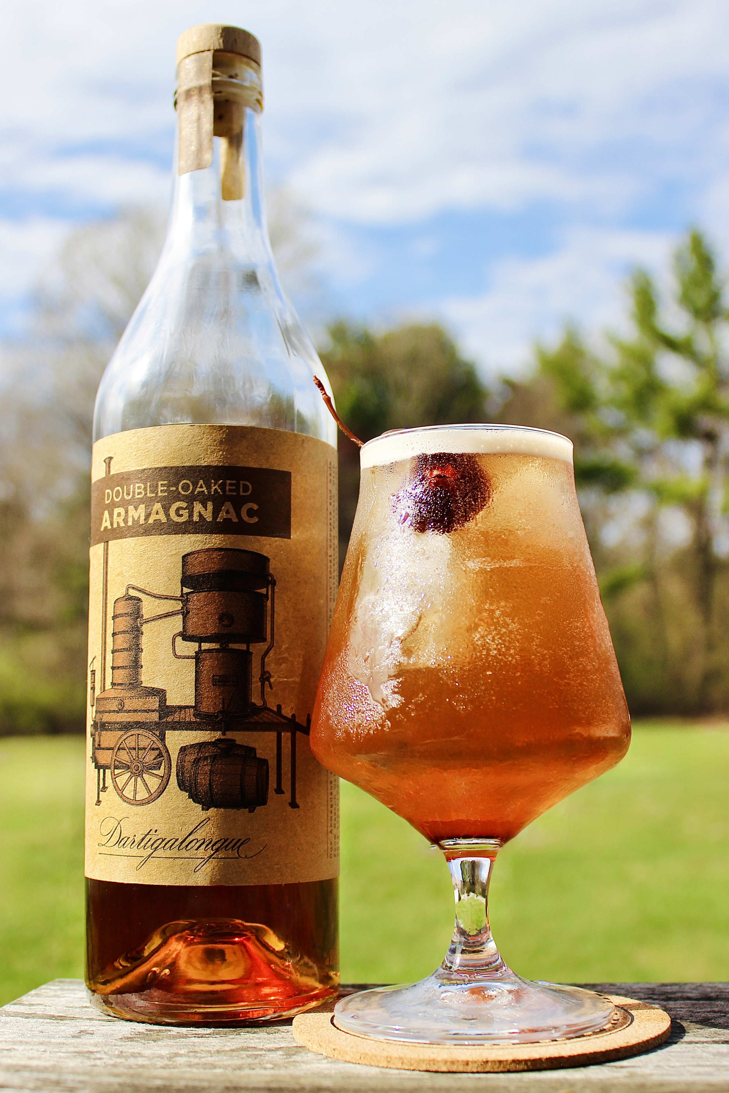 A stem glass holds a bubbly cola-colored cocktail topped with foam and a brandied cherry. Next to it is a bottle of Dartigalongue Double-Oaked Armagnac against a bright, sunny outdoor backdrop.
