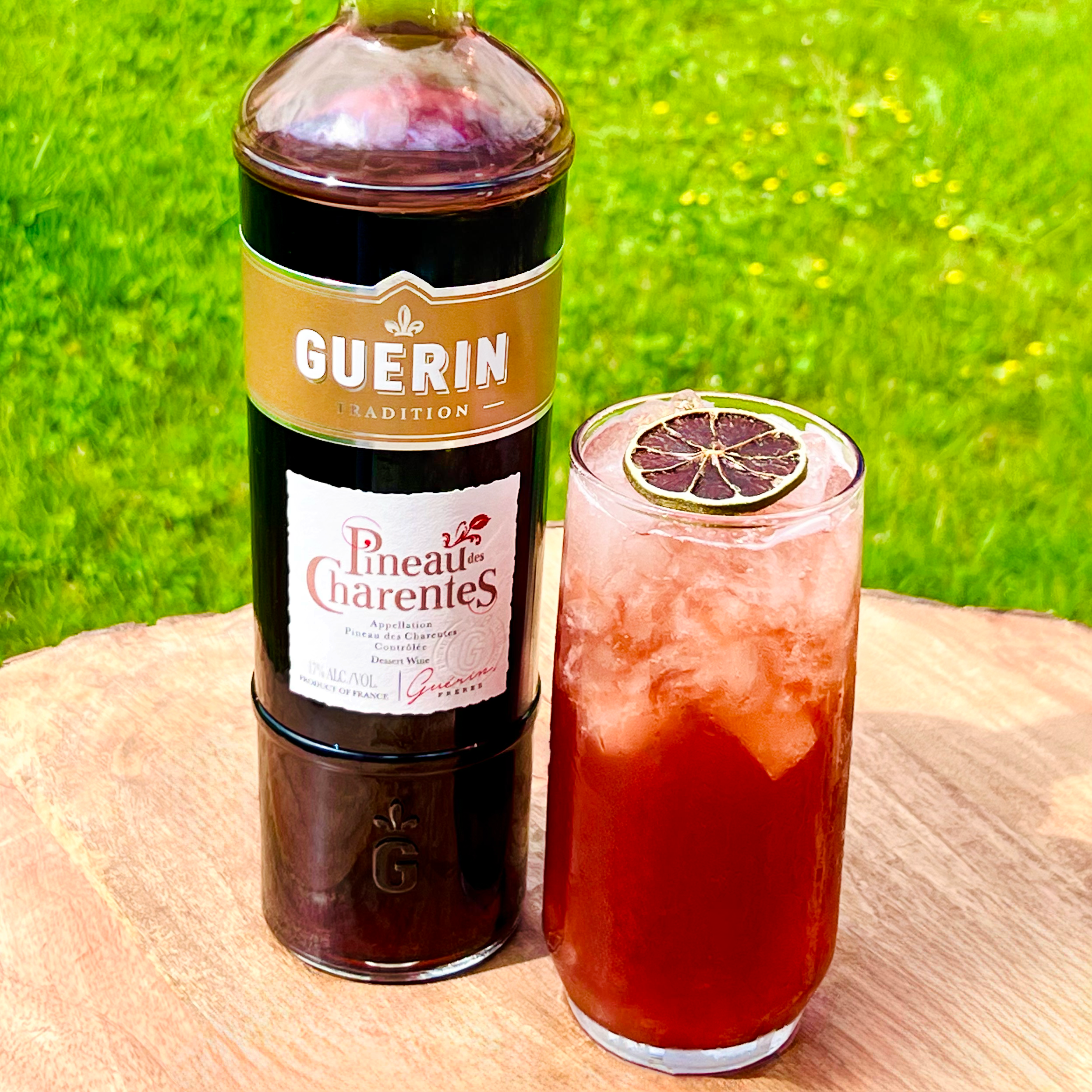 A bottle of Guérin Tradition Pineau des Charentes next to a tall glass of pink cocktail with ice and a dried lemon slice on top, placed on a wooden surface outdoors with green grass in the background.