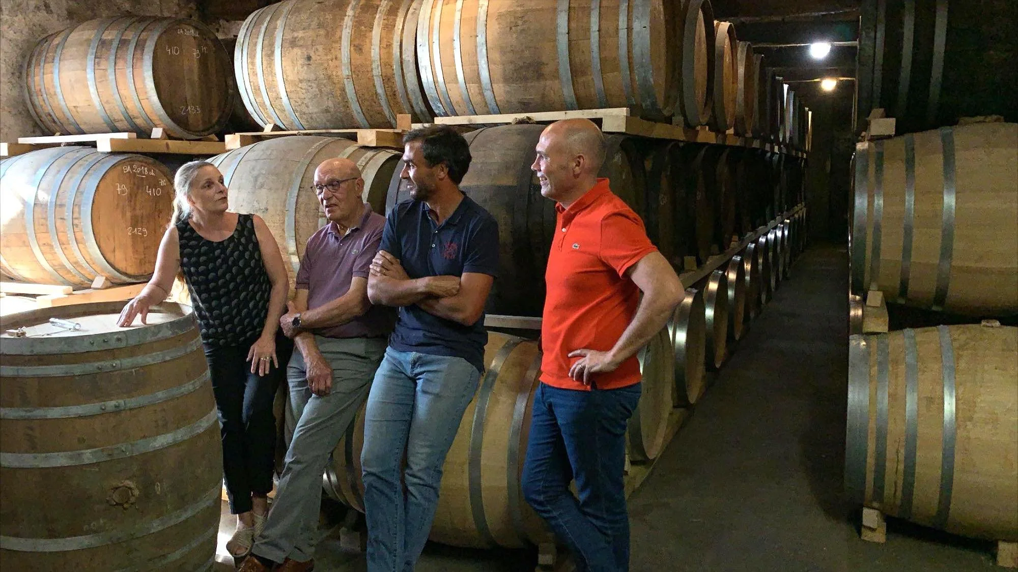 A woman stands next to a man and his two adult sons, chatting inside a dimly lit cellar lined with stacked oak barrels at Armagnac Delord. They appear engaged in conversation, surrounded by rows of aging barrels.