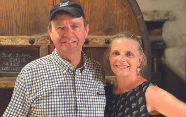 Man in baseball cap and woman stand in front of large wooden calvados barrel