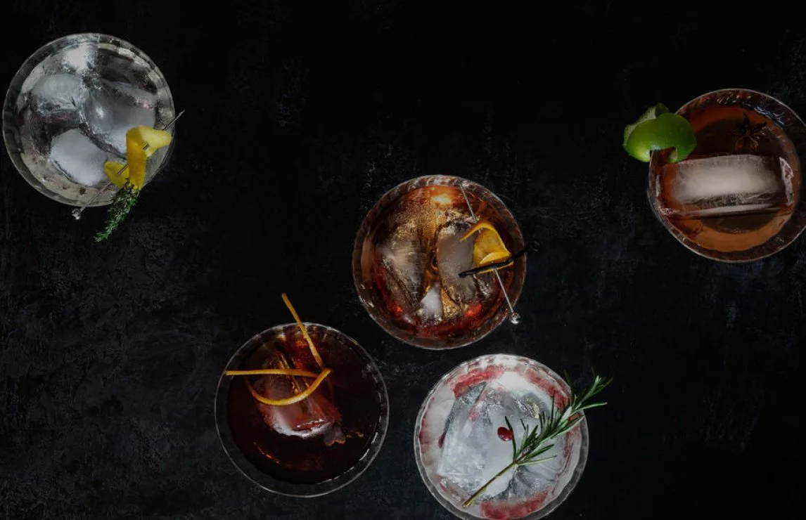 Overhead view of several cocktails with ice in glasses on a black surface. 