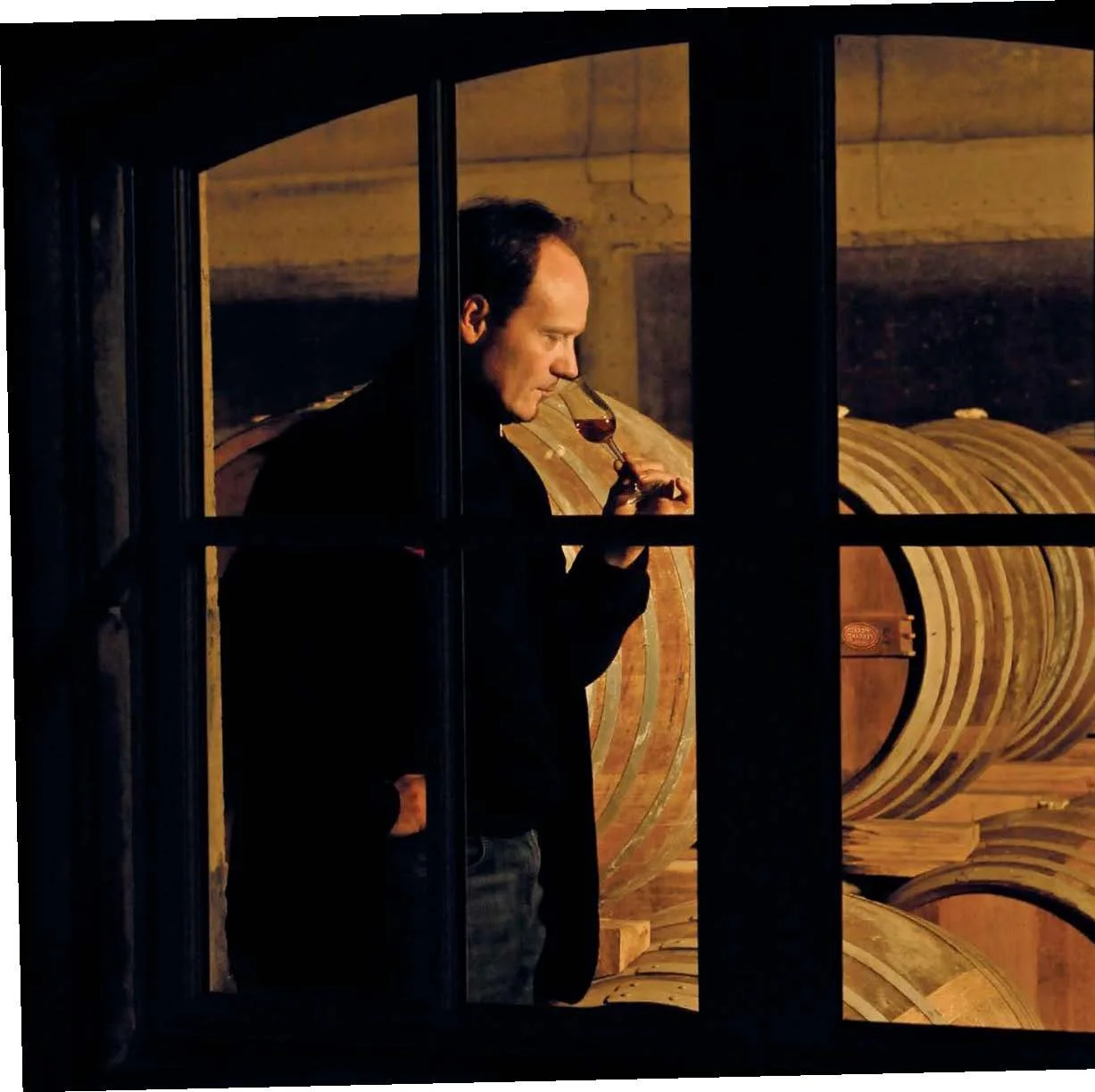 Man in a brandy cellar smelling a glass of cognac as seen through a window