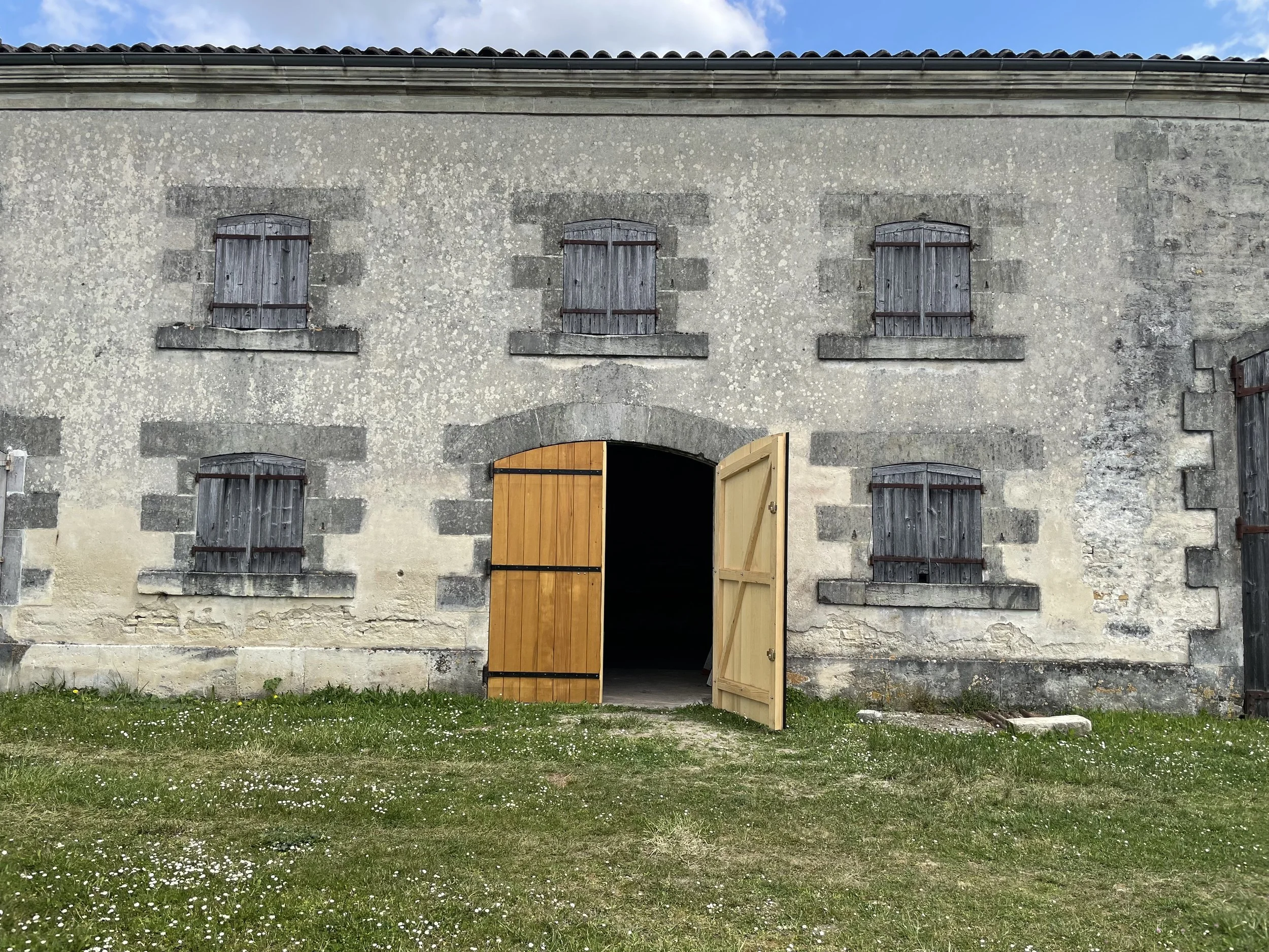 Pale stone building with wooden shutters and darker stone lintels with a double honey-colored arched wooden door with one door opening into darkness.